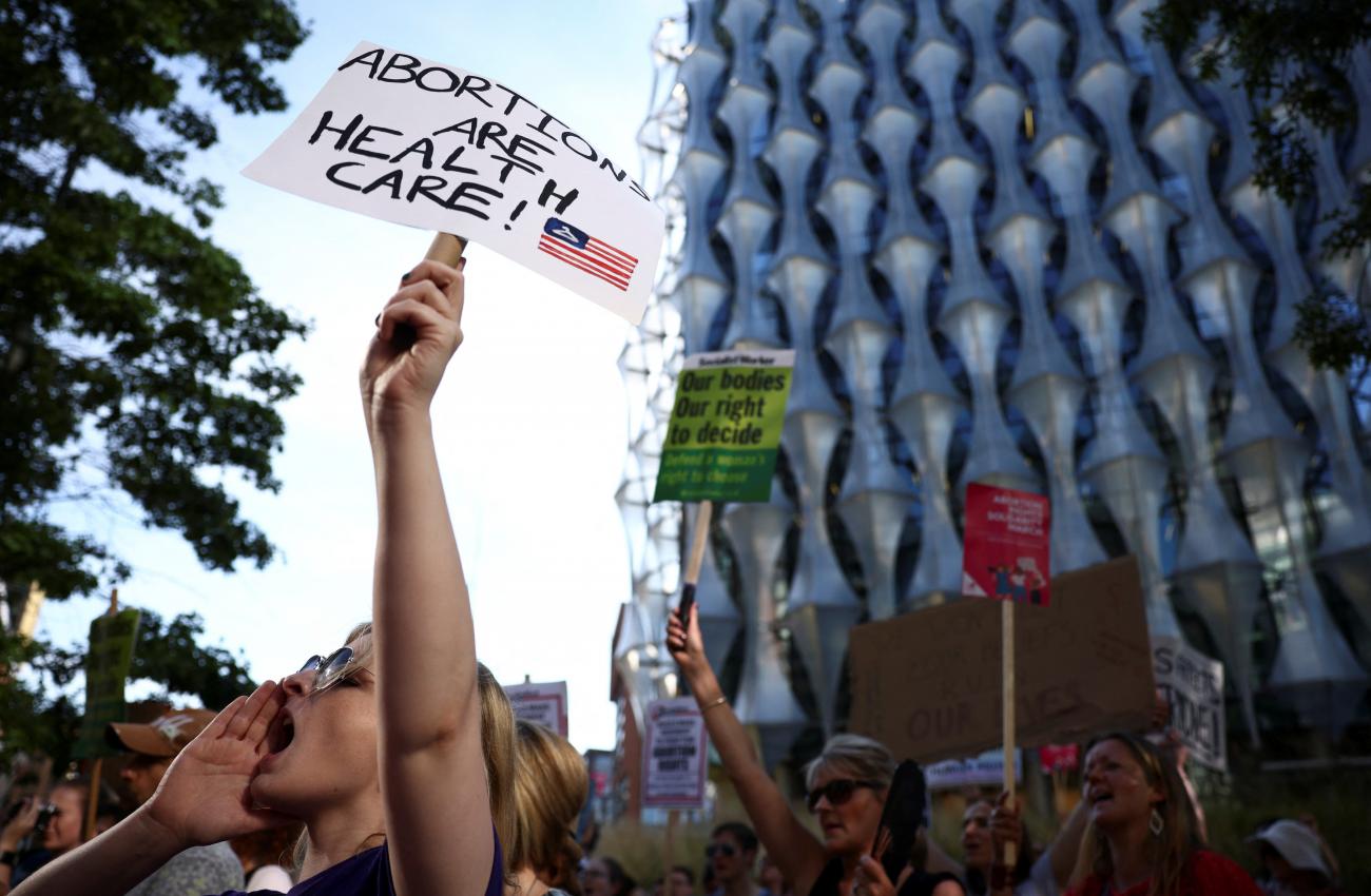 A blonde woman stands in front of a crowd at a protest yelling with one hand against her mouth and the other extended holding a white poster-board sign that reads "abortions are health care" in dark blue marker