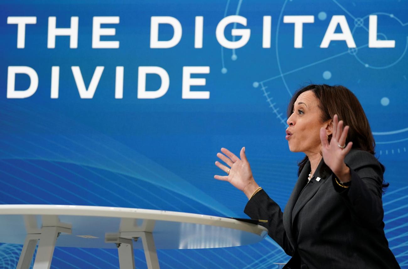 U.S. Vice President Kamala Harris is photographed from the side wearing a dark gray suit a gesturing with her hands in front of a blue screen that reads "the digital divide" in white letters