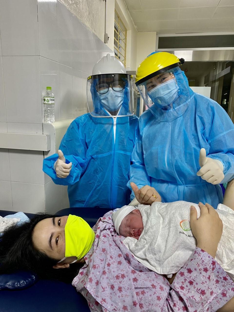 A woman in a bright yellow mask smiles as she holds her newborn infant to her chest. Behind her, two medical professionals in blue medical smocks and masks give a thumbs up and smile.