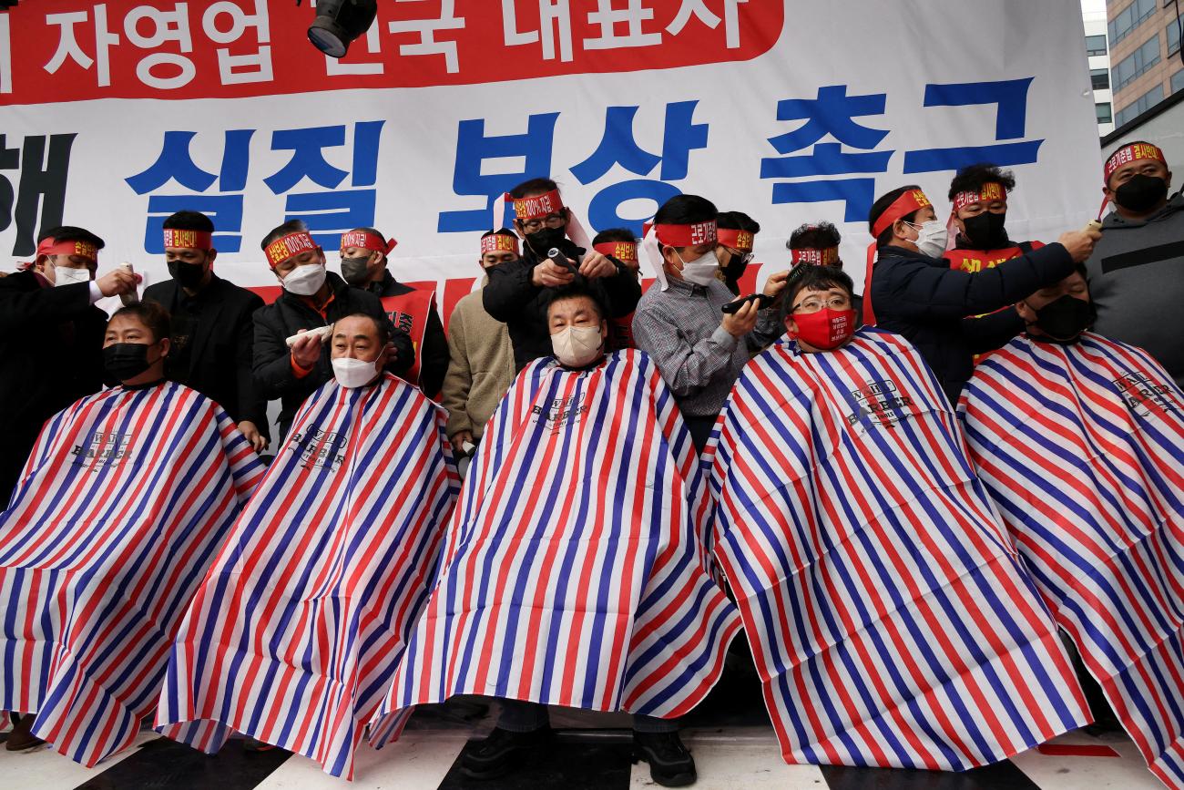Five business owners sit in a row in barbershop chairs, wearing colorful smocks, having their heads shaved to protest against the government's new social distancing rules, in Seoul, South Korea, on January 25, 2022.