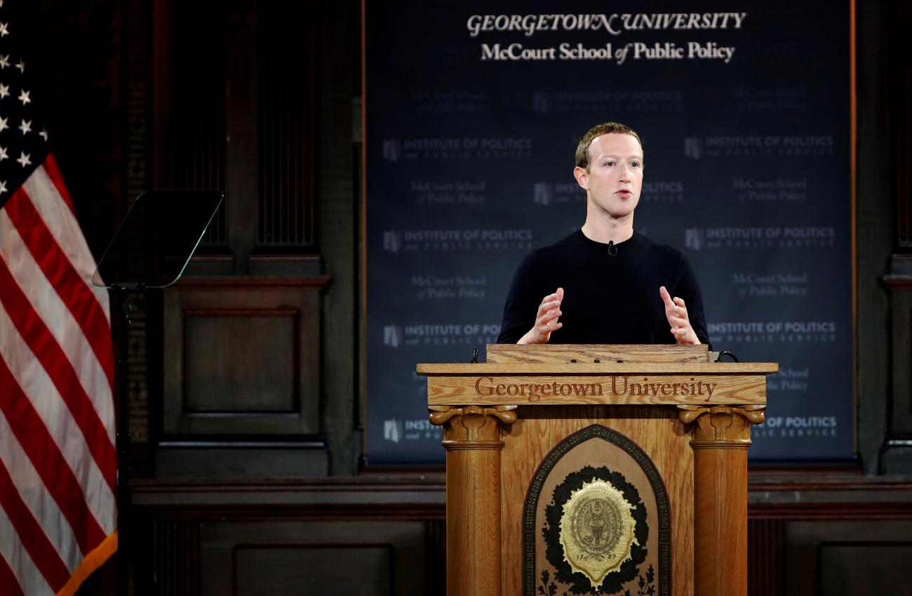 Facebook CEO Mark Zuckerberg stands at an ornate wooden podium next to an American flag and gesticulates as he addresses an audience in front of a blue background that says "Georgetown University McCourt School of Public Policy"