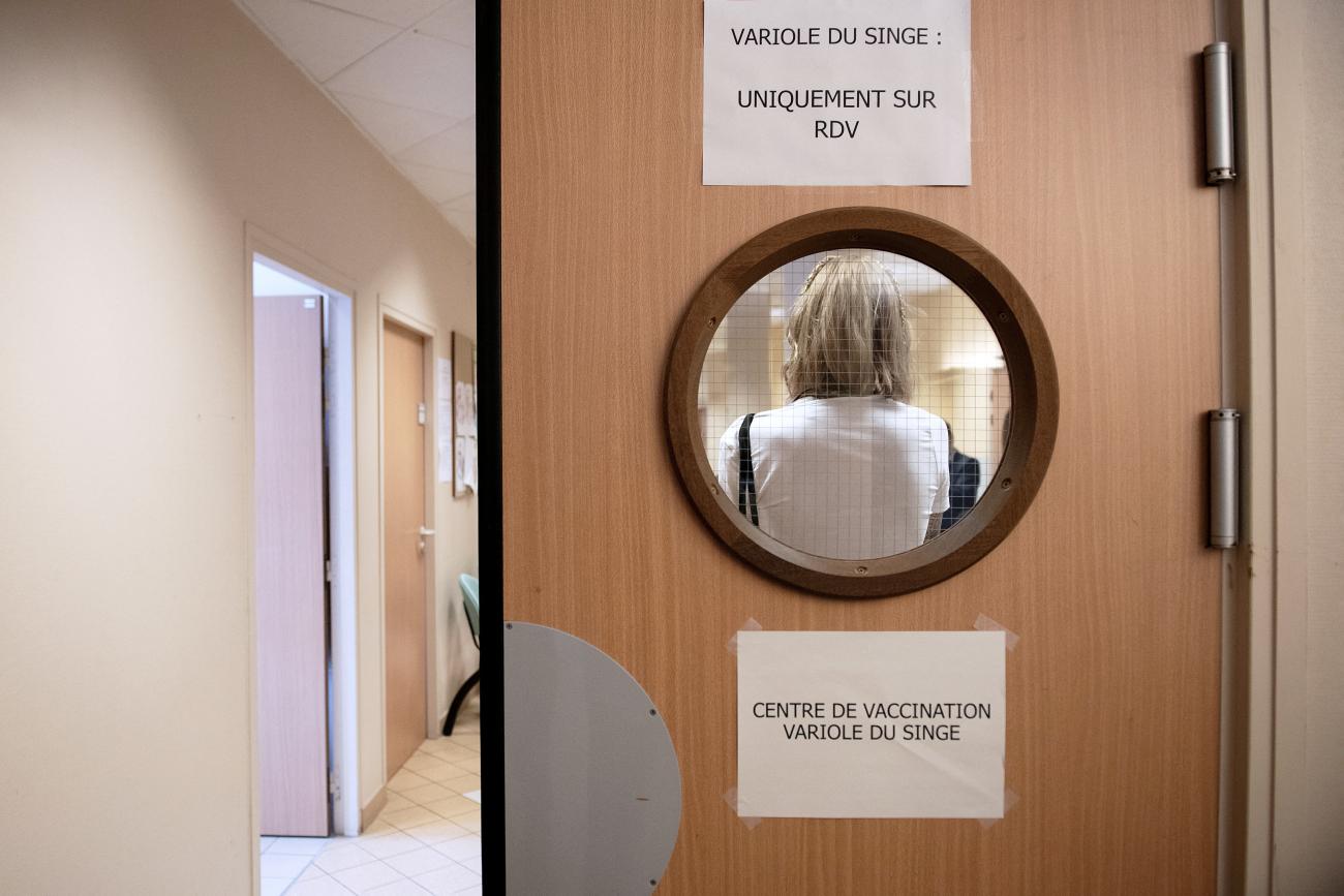 The back of a blonde woman's head is photographed through a small circular window cut out of a light-oak hospital door