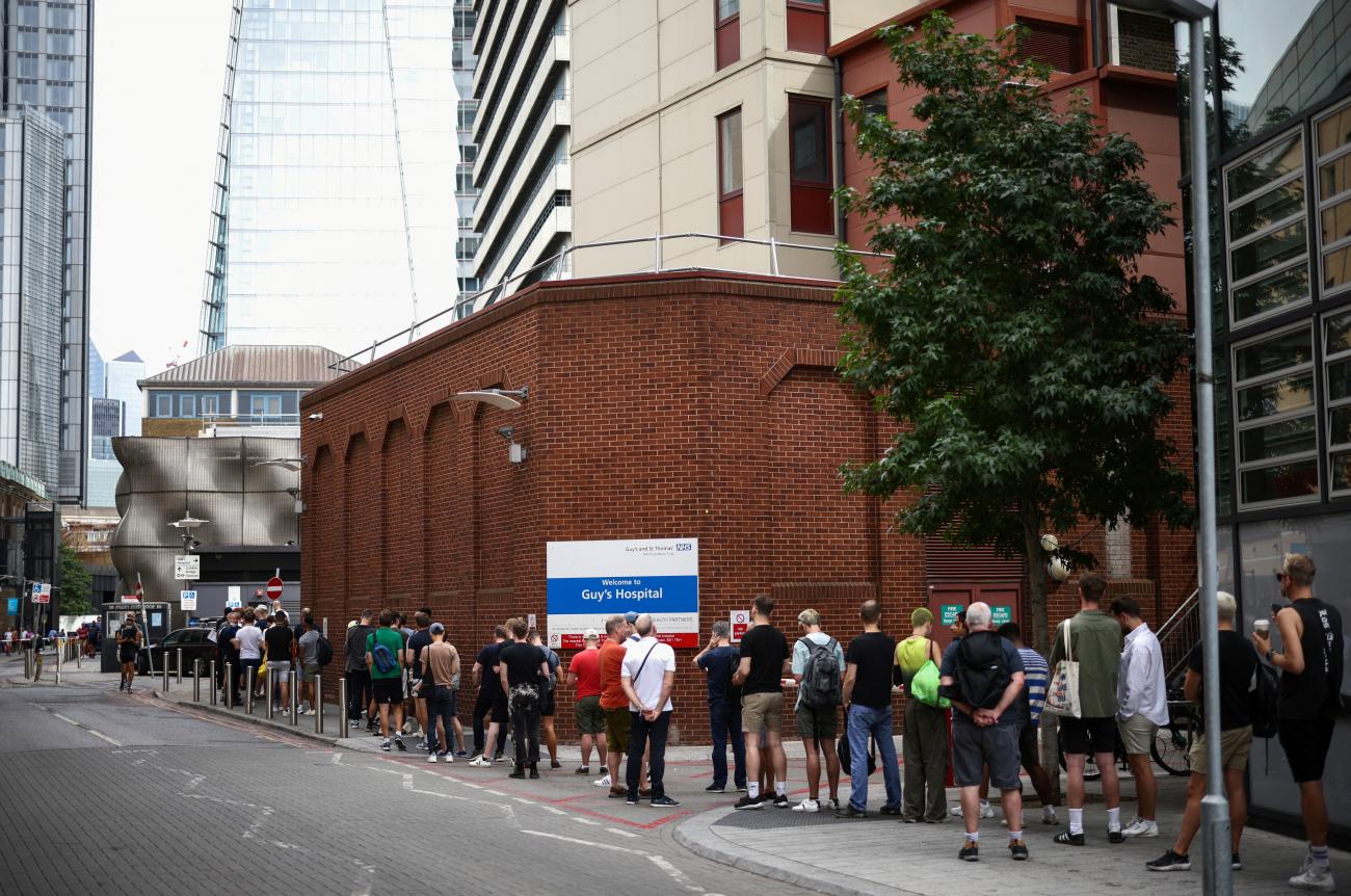 Men wearing summer clothing queue up in front of a red brick building with a blue sign that says Guy's Hospital. In the background London skyscrapers glint in the sun. 