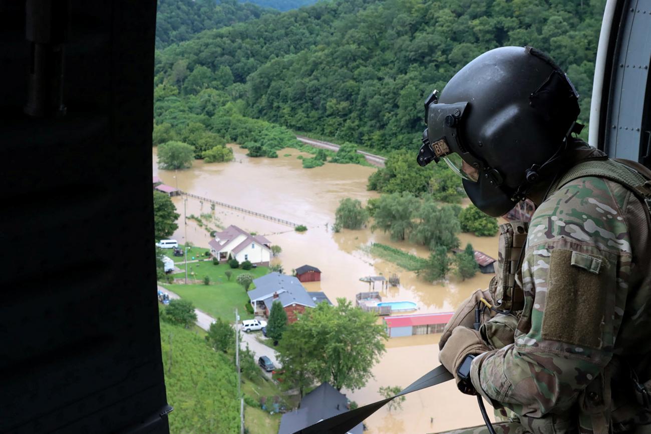 A Kentucky National Guardsman in green camouflage and a black helmet looks out of the open door of a helicopter onto a flooded landscape in which houses and swimming pools can be seen partially submerged in muddy water.  