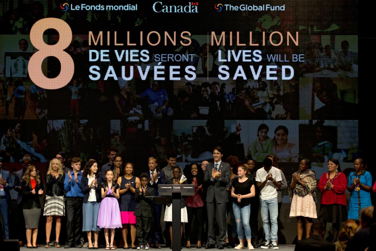 Prime Minister Justin Trudeau stands among conference attendees against a mosaic backdrop with projected text that reads "8 million lives will be saved"