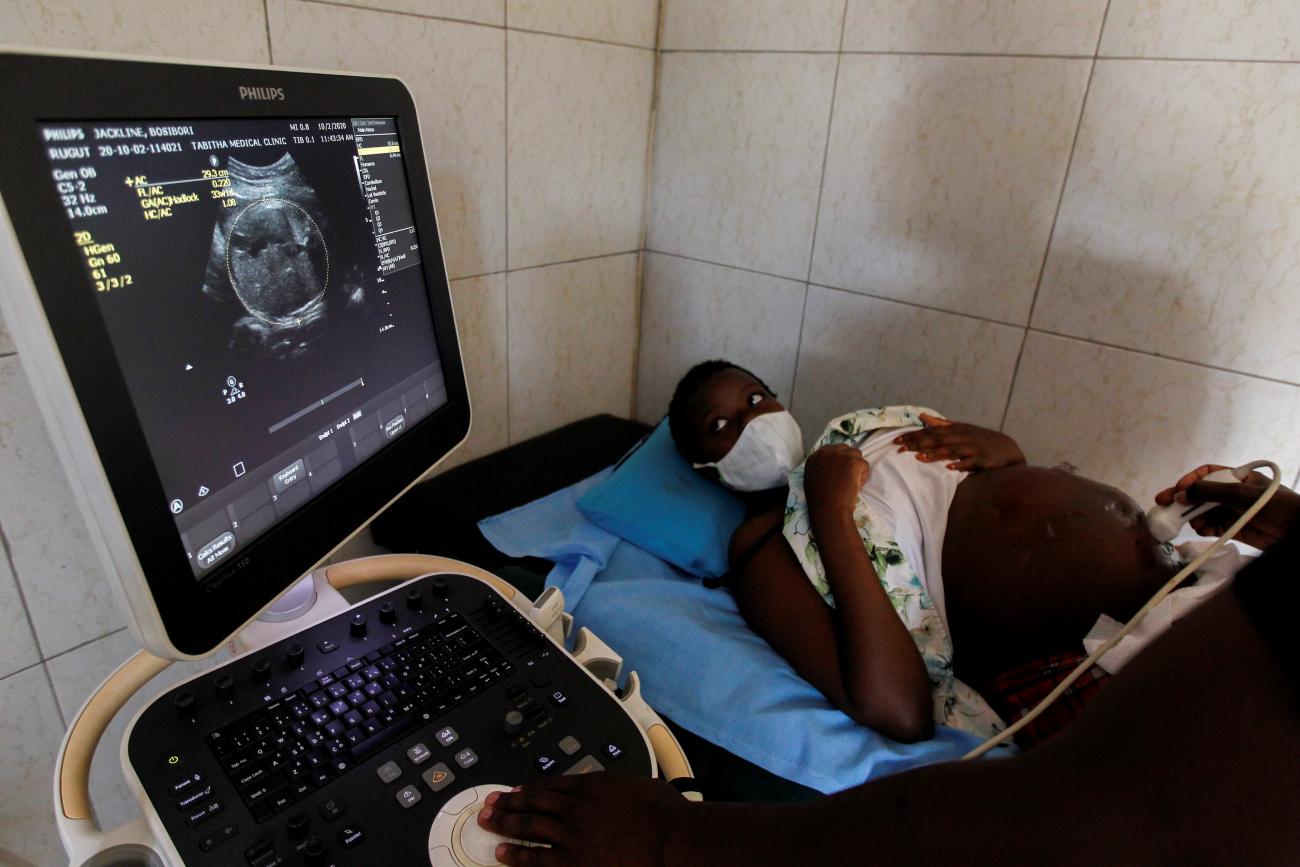 A young pregnant woman wearing a white surgical facemask lays down on a hospital bed with her hands placed over her chest as she looks onto an ultrasound screen