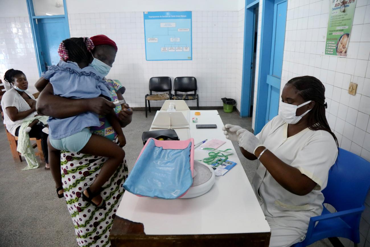 A nurse wearing white scrubs and a white surgical facemask prepares a malaria vaccine dose for a baby being held by its moter