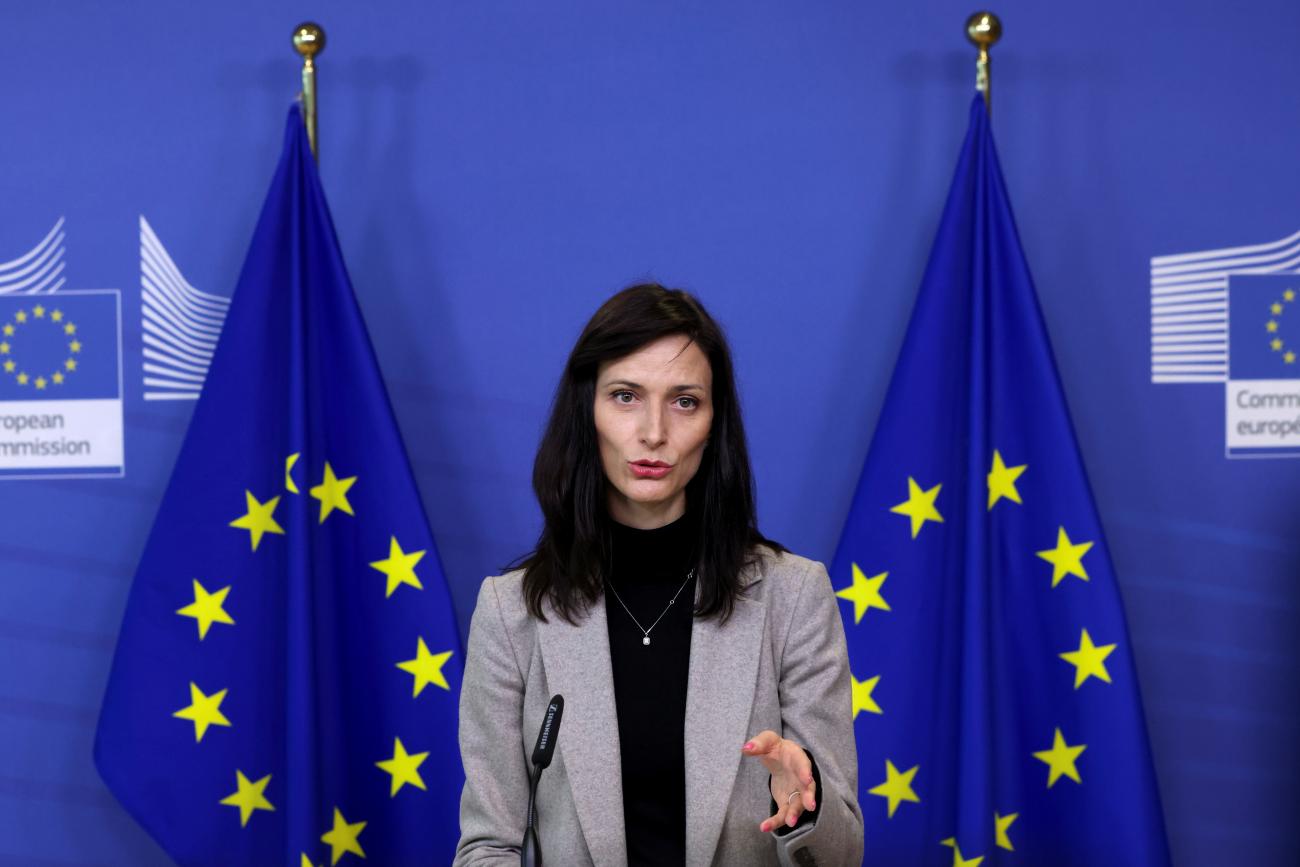 European Commissioner official Mariya Gabriel, who has long dark hair and is wearing a grey blazer, stands in front of a dark blue background as she speaks at a news conference. On either side of her are the blue flags with yellow stars of the European Union.