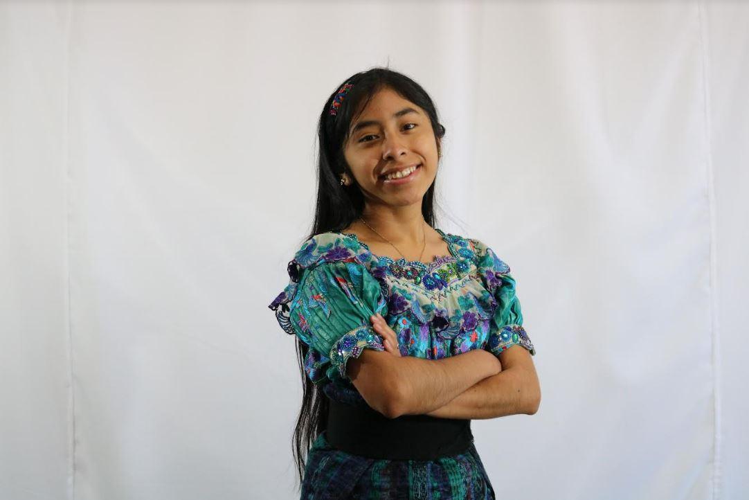 For her Huertos Familiares project, Marisol, 17, teaches local families how to plant backyard gardens. Marisol is pictured smiling and with her arms crosses. She has long brown hair and wears a green blouse with embroidery, at MAIA Impact School in Sololá, Guatemala, 2022.