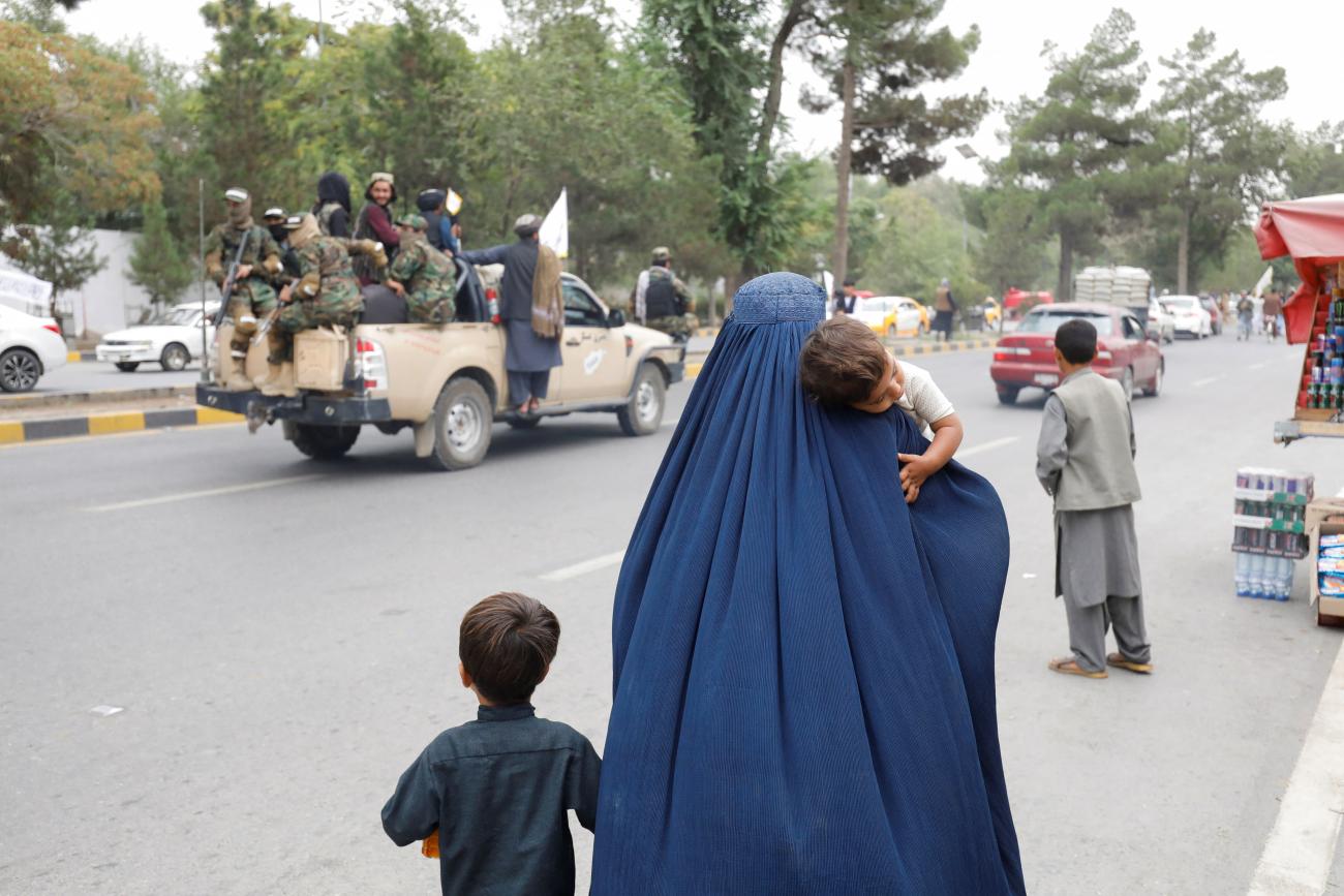 An Afghan woman in a blue burqa holds one of her children by the hand and carries the other on the anniversary of the fall of Kabul. In the background a truck full of Taliban soldiers drives by. 