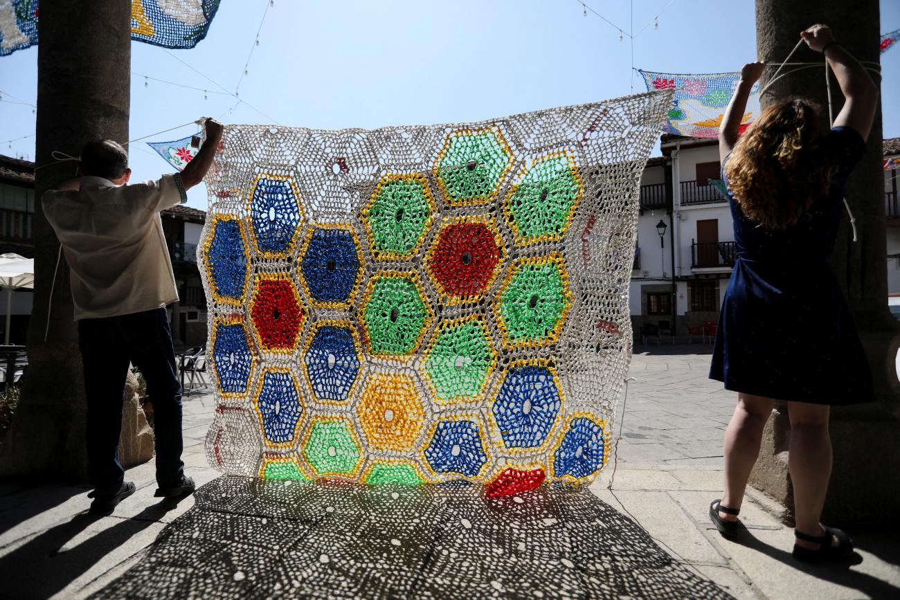 Volunteers, silhouetted by the afternoon sun, hang a colorful tapestry woven of white, blue, red, and green recycled material above a pedestrian pathway. In the background, other colorful tapestries are already shading the cobbled streets below.