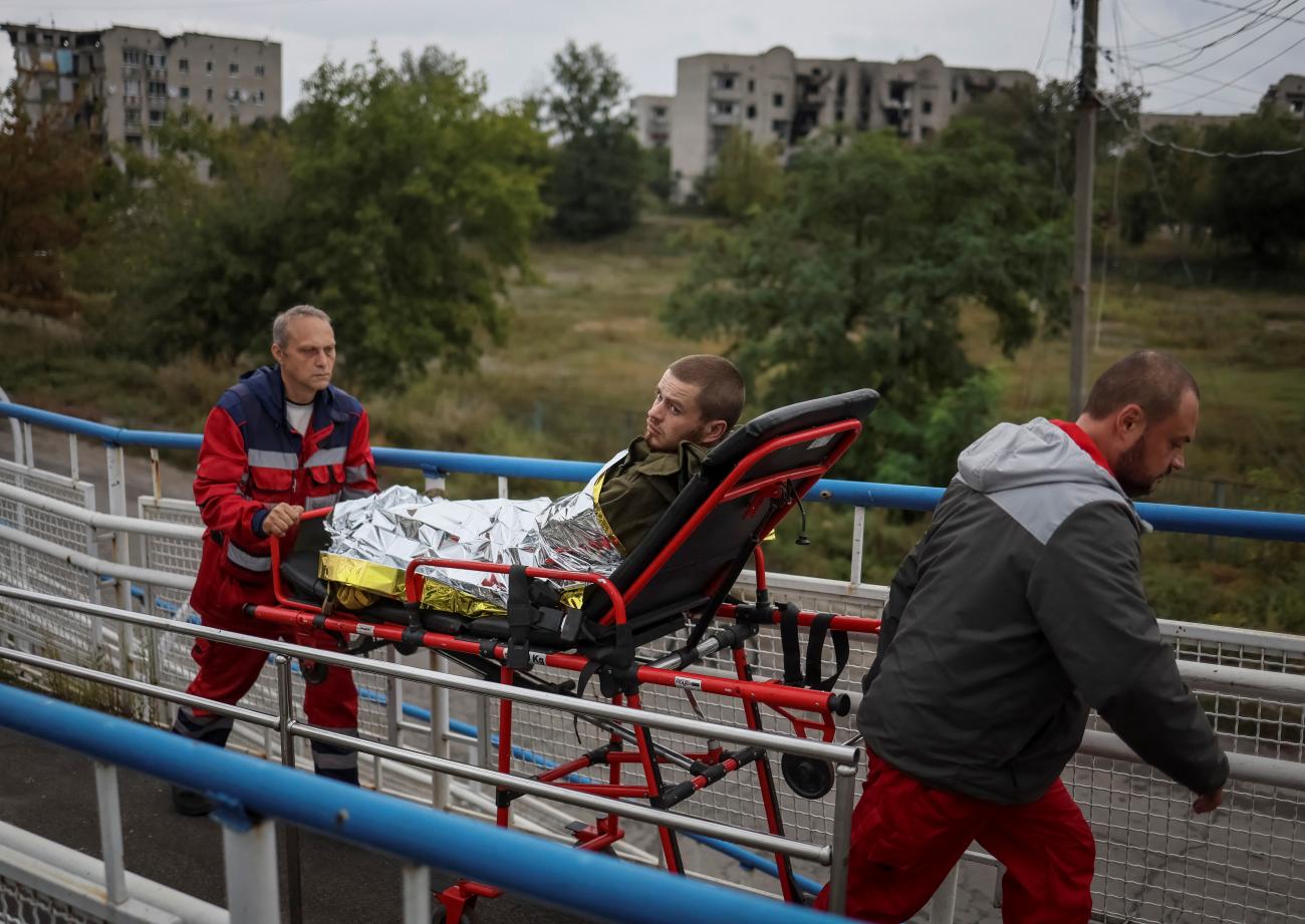 Two health workers in red emergency outfits transport a wounded Ukrainian serviceman on a stretcher in the town of Izyum, recently liberated by Ukrainian Armed Forces. In the background, are trees and concrete apartment buildings that have been damaged by shelling. 