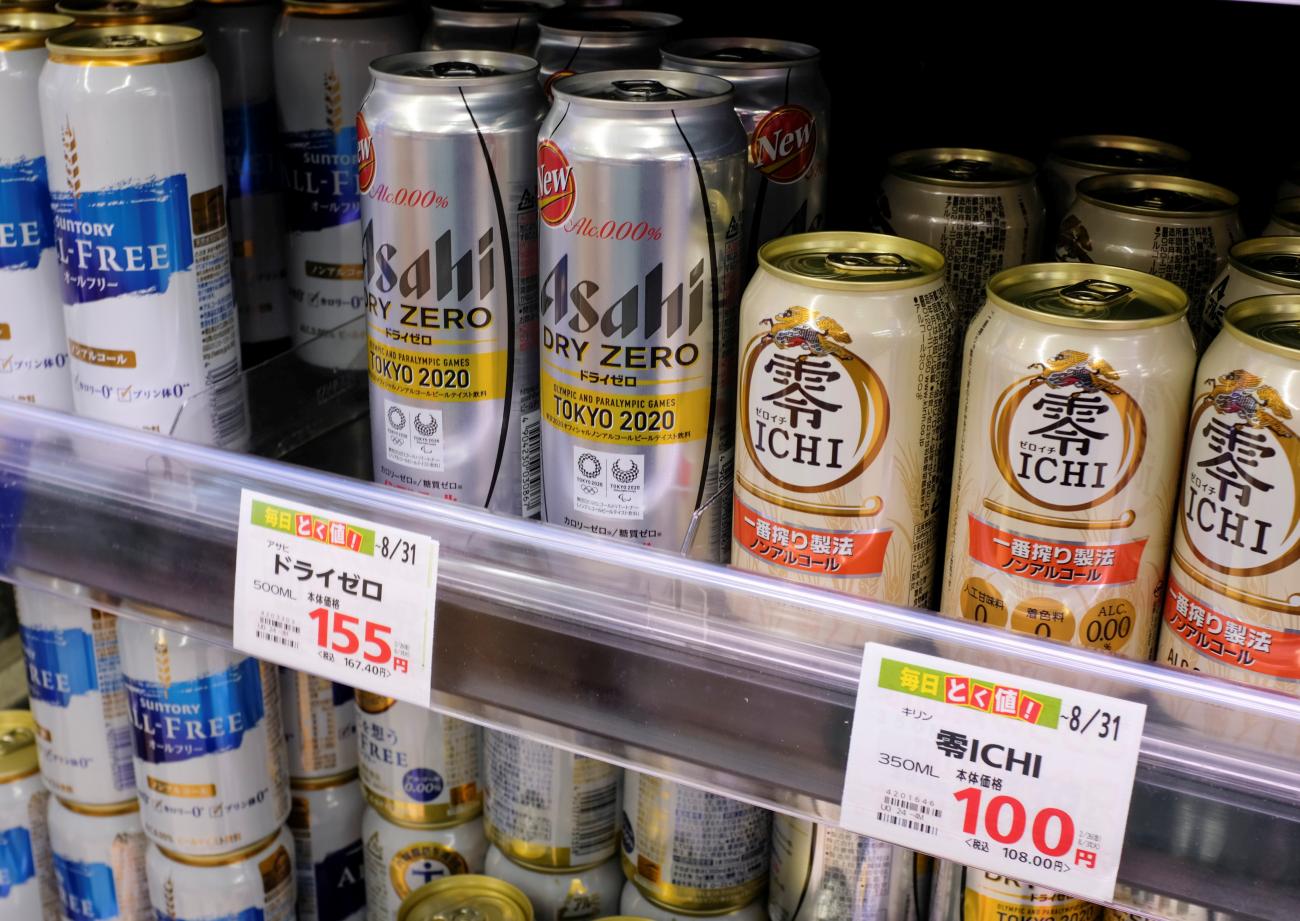 Various cans of alcohol-free beers, including those from Asahi, Kirin, and Suntory, are displayed on a supermarket shelf in Tokyo, Japan. White price tags with orange letters show prices of 100 yen for a small can and 150 yen for a large one.