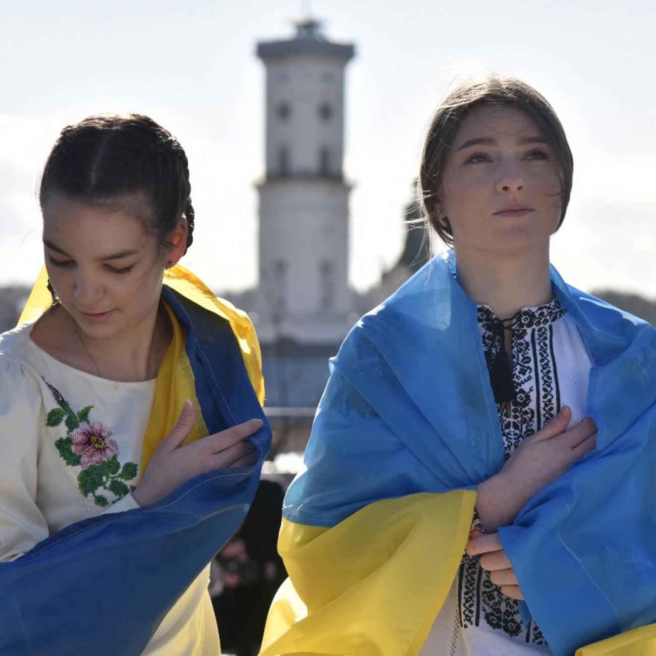Two girls stand in Lviv, Ukraine wearing traditional Ukrainian blouses and wrapped in the blue and yellow Ukrainian flag