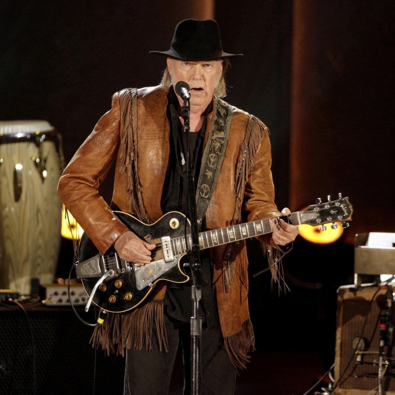 Singer-songwriter Neil Young performs on stage wearing a fringed jacket and black hat.