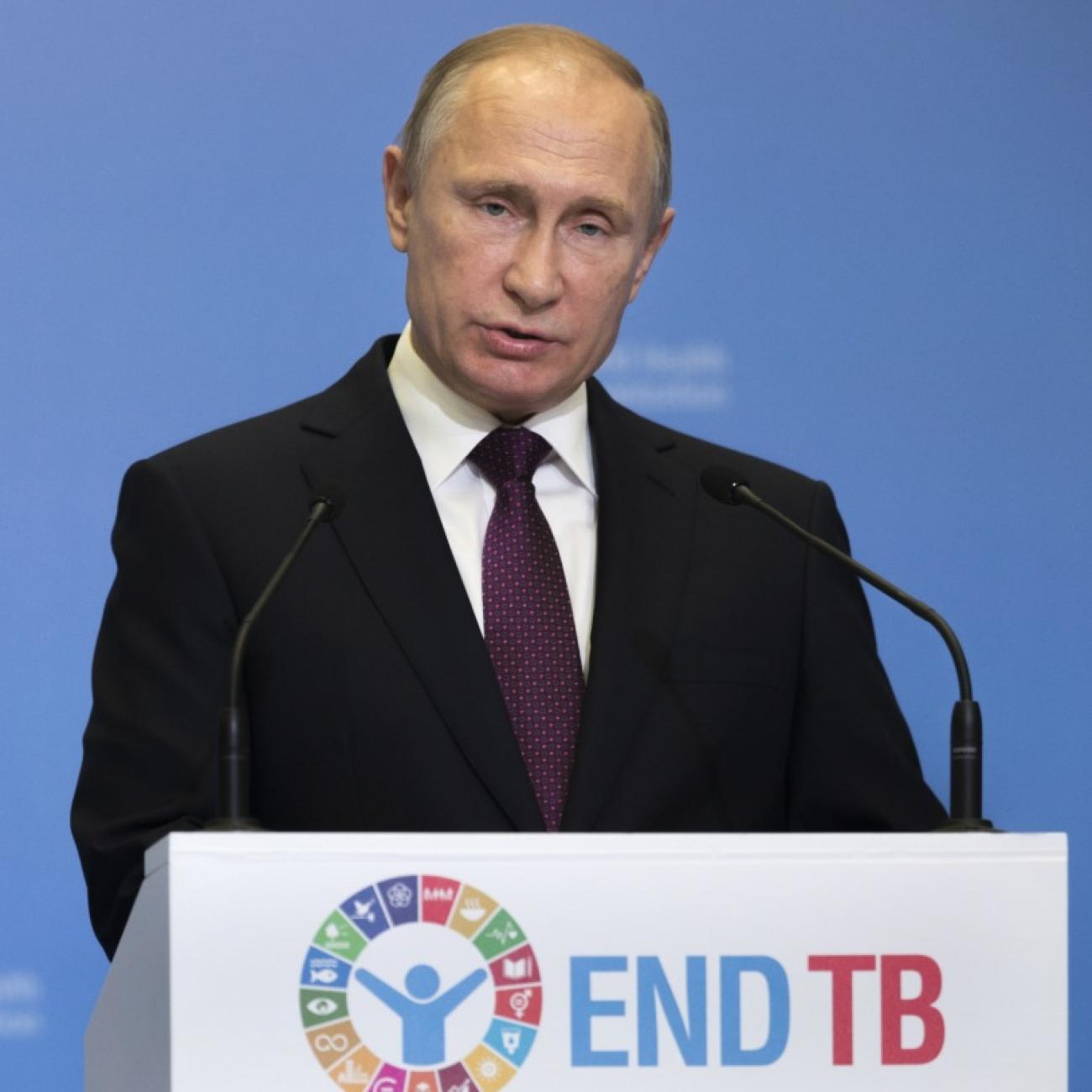 Russian president Vladimir Putin stands in front of a podium that says "End TB" with a bored expression on his face.  