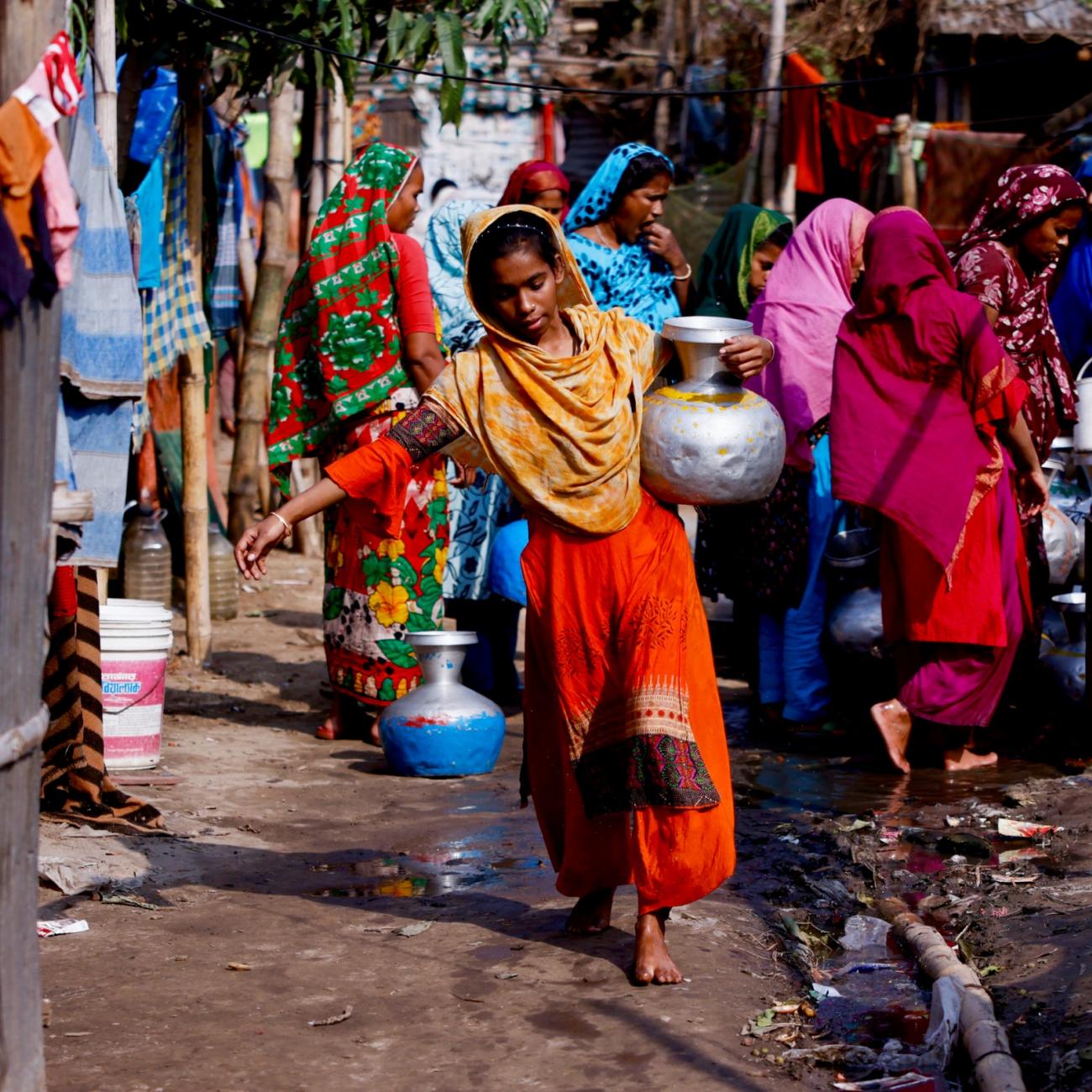 A woman in an orange dress carries a water jar after collecting water from a common water tap. In the background are other women in colorful garments who don't have direct access to water at home in Dhaka, Bangladesh, February 21, 2022. 