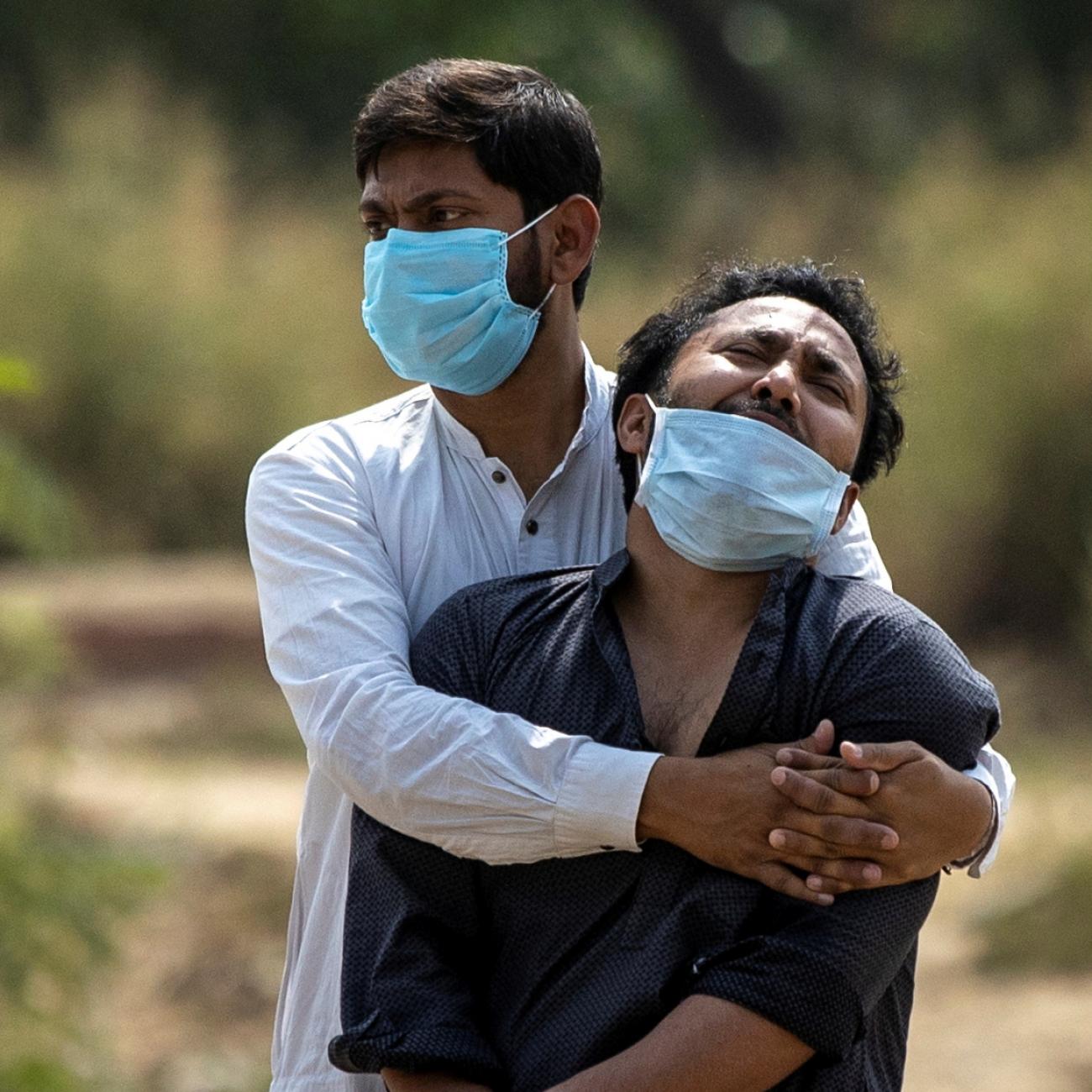 A young man in a white shirt wearing a blue surgical mask hugs his brother, wearing navy blue, as he wails mourning the loss of his father from COVID-19
