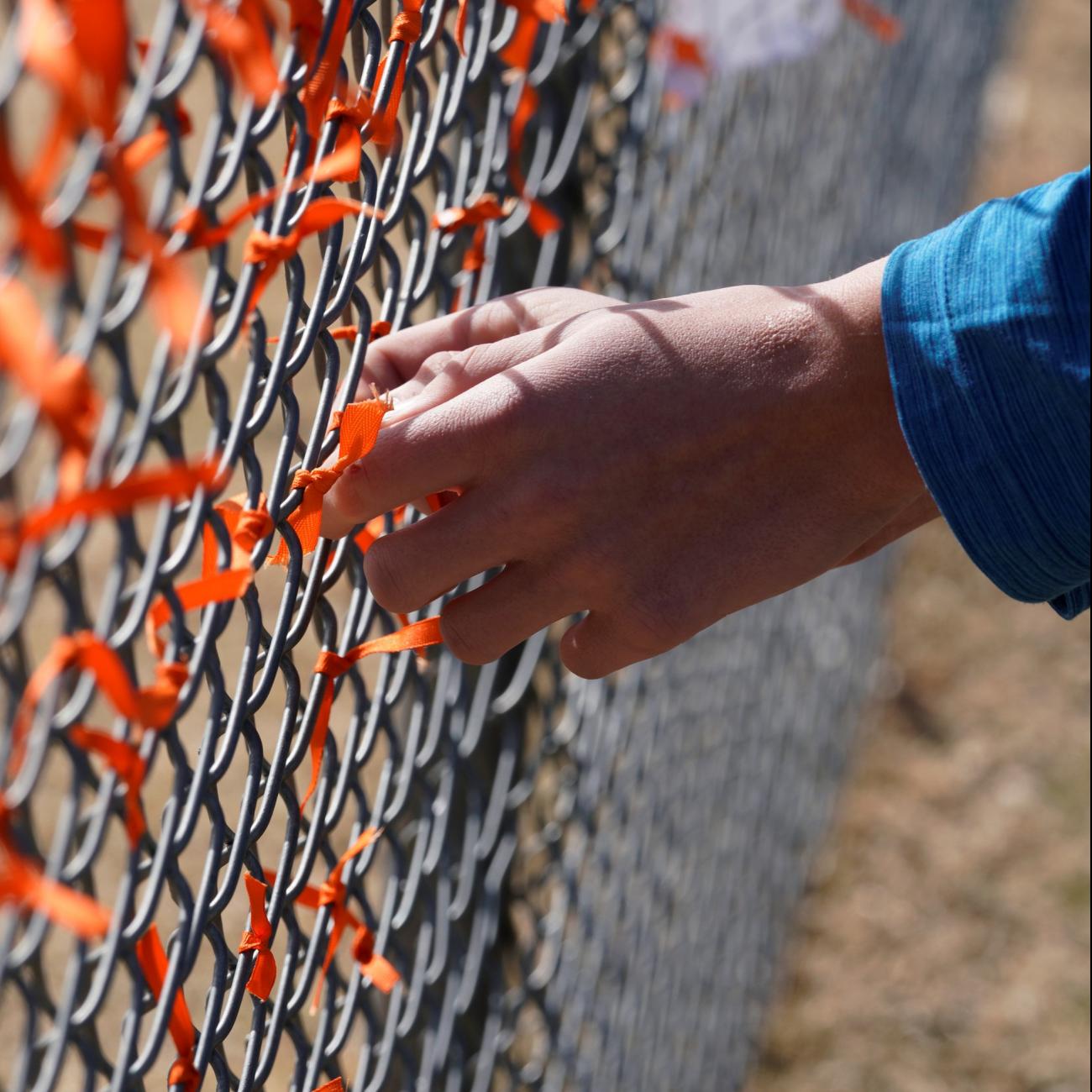 A white hand with the cuff of a blue jacket reaches out to tie an orange ribbon onto a chain-link fence covered in orange ribbons in honor of those killed by gun violence