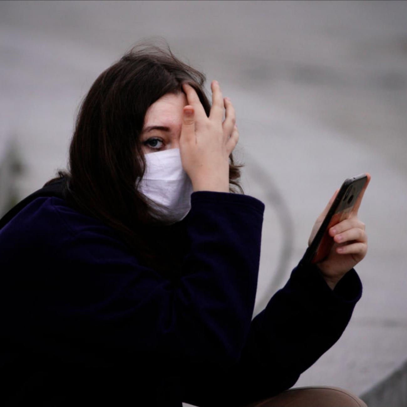 A young woman with dark hair in a black coat and white mask holds a black phone in one hand and covers part of her face with the other. She looks serious as she sits on grey stone steps in Warsaw, Poland.