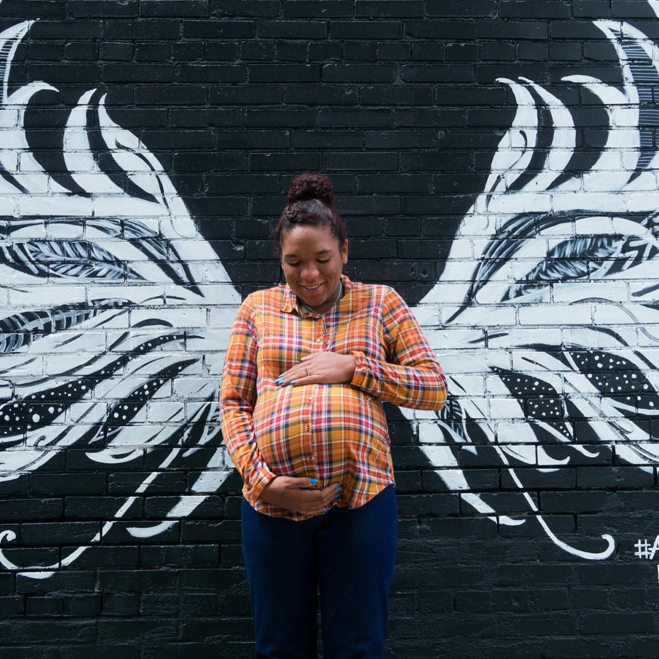 A Black mother in an orange shirt lovingly holds her pregnant belly in front of a mural on a black brick wall that gives her white angel wings in Anna, Illinois, U.S., May 10, 2021.