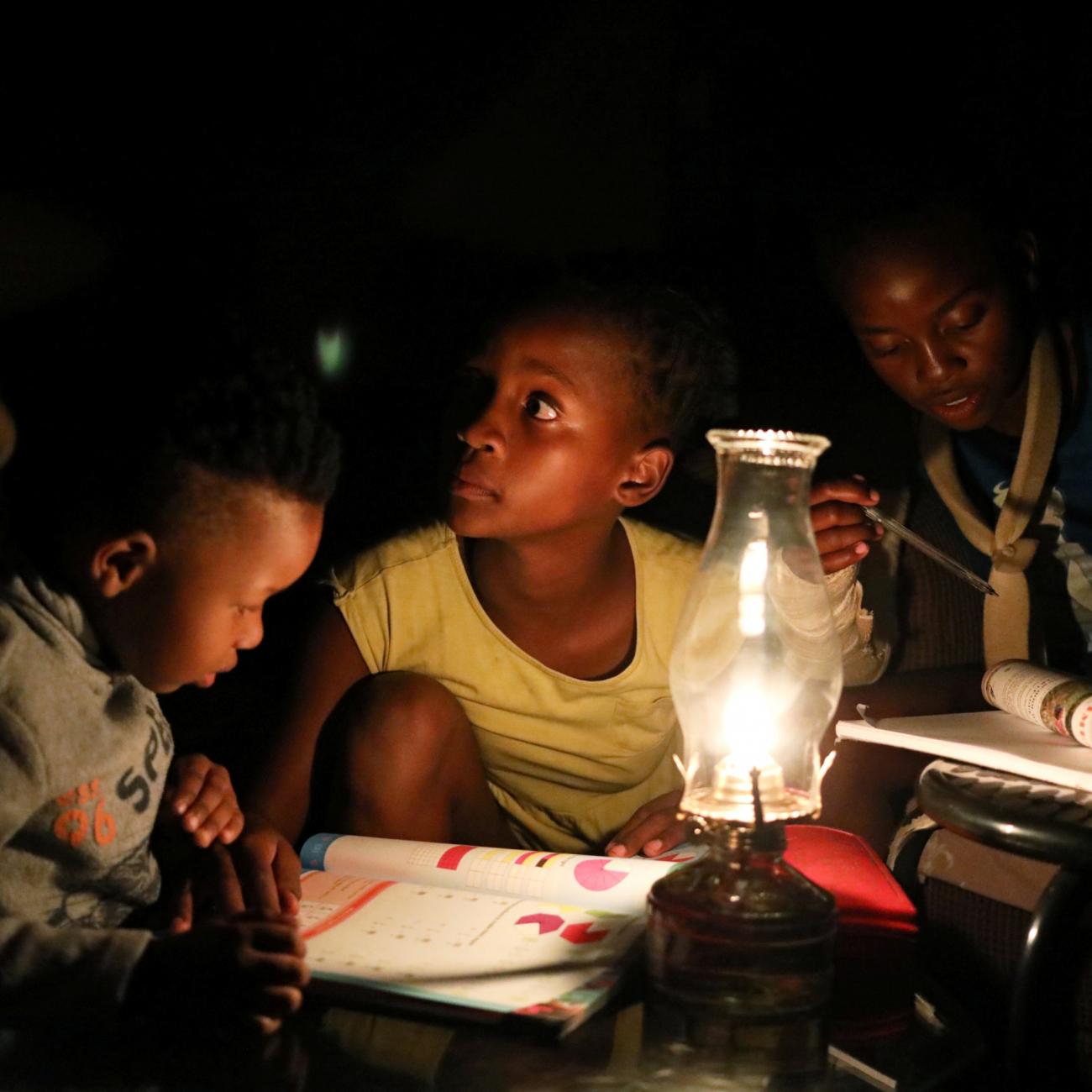 Three South African children sit in a dark room look on as they use a parafin light to study during an electricity load-shedding blackout in Soweto, South Africa, on March 18, 2021. 
