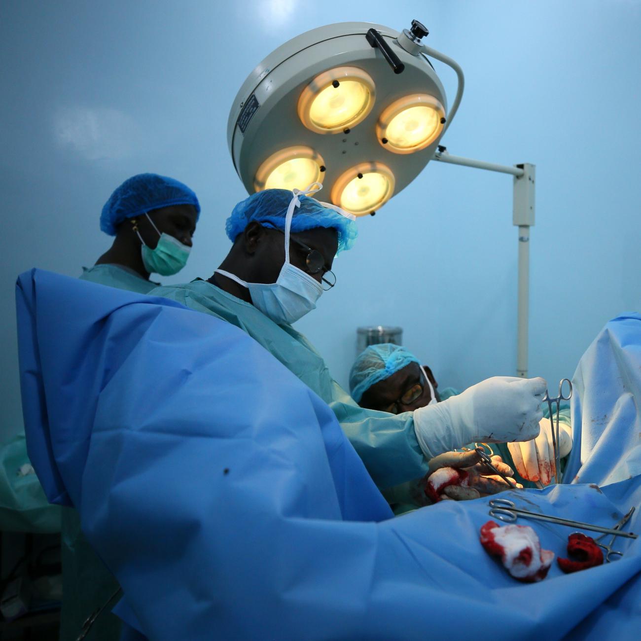 Nigerian surgeons in blue gowns and masks operate on a patient hidden beneath a blue sheet under a large surgical lamp in a room with blue walls. 