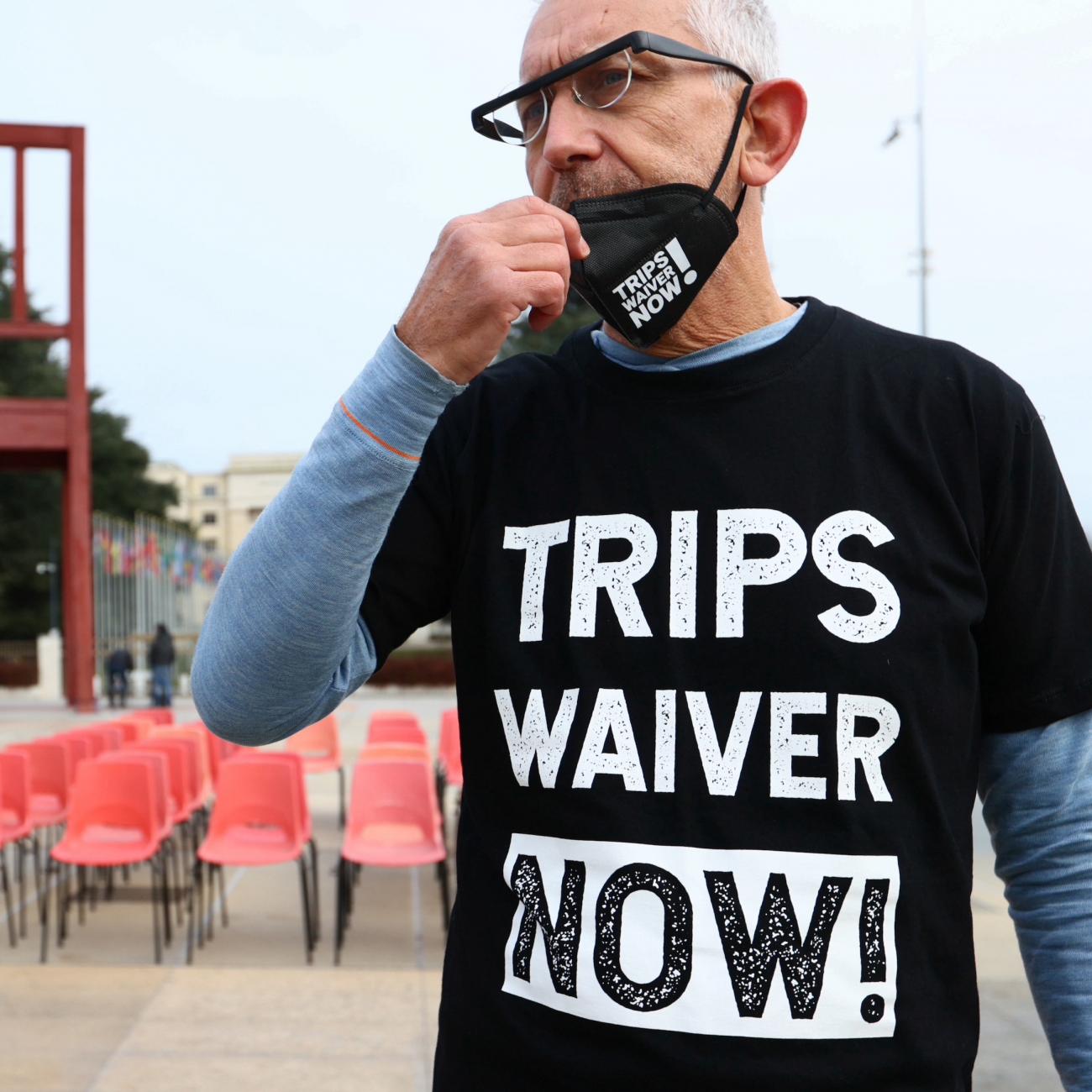 A man adjusts a face mask and is wearing a black shirt that reads "TRIPS WAIVER NOW!" He stands in front of rows of empty chairs.