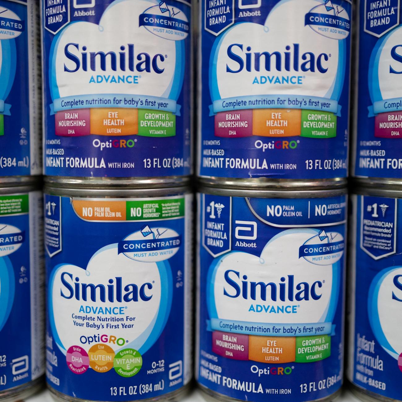 Eight blue cans of baby formula with white, orange, and green labels that read "Similac" stand in stacks of two on a pharmacy shelf.