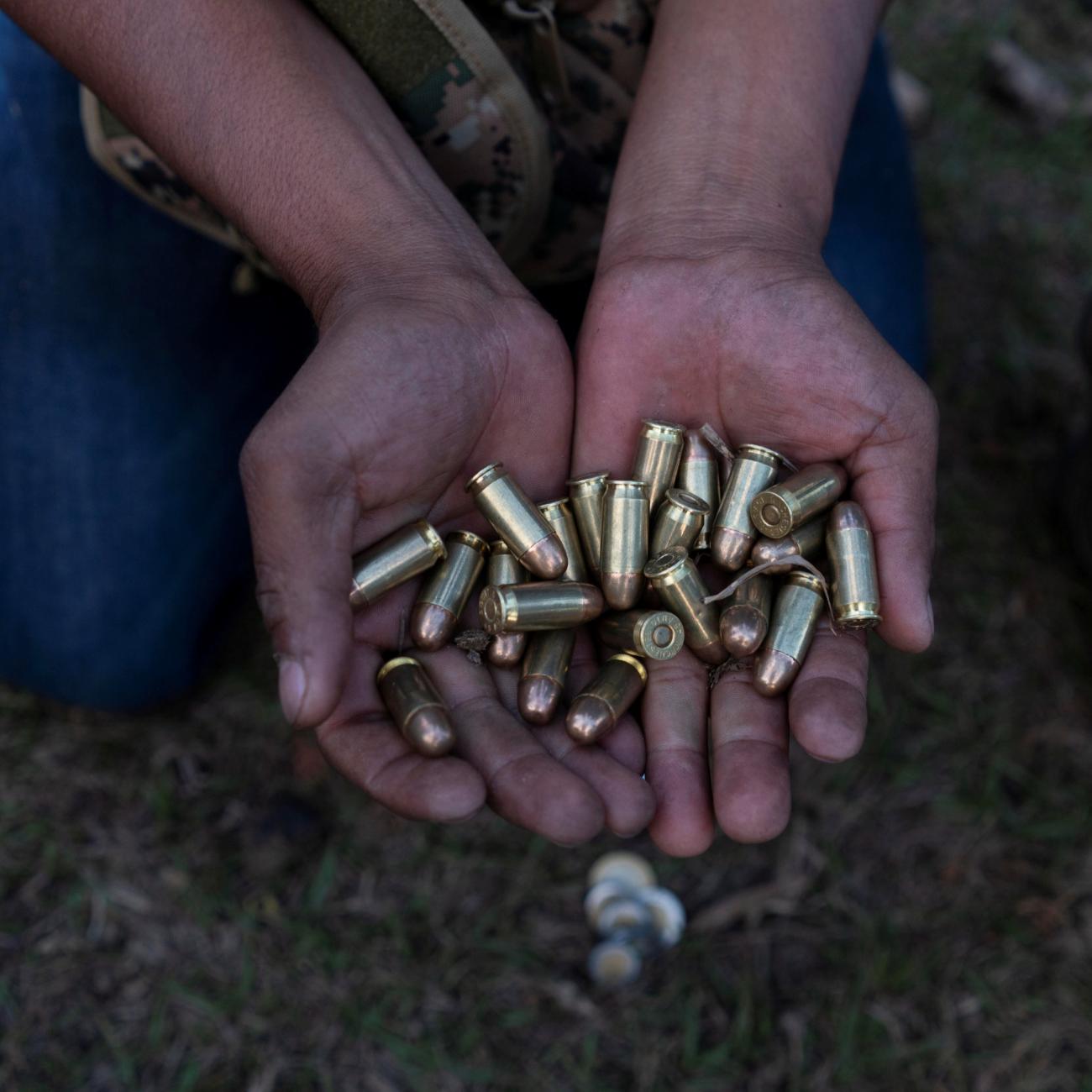 A community police officer in blue jeans squats low to the ground and holds out two hands full of shiny metal 45mm caliber bullets