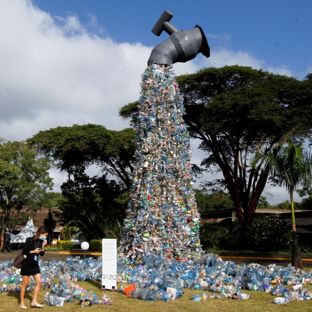 A female delegate wearing a long sleeved black dress a blue surgical mask looks at a 30-foot sculpture made out of plastic bottles resembling a faucet, outside the UN Environment Programme Headquarters, in Nairobi, Kenya 