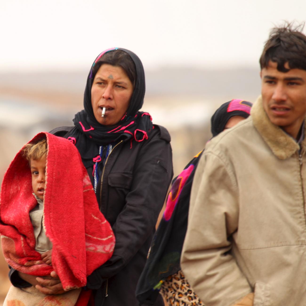 An Syrian woman with a serious expression on her face dressed in dark colors holds a child who is partially obscured by a bright orange scarf draped over his head. On either side of her, are young men in winter coats walking with her through a desert landscape