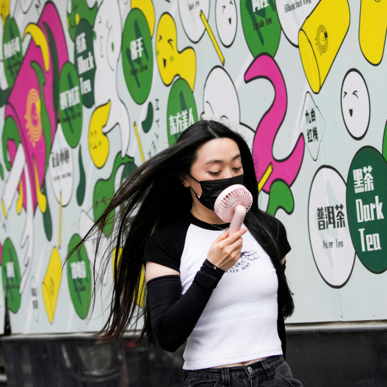 A woman in a white and black t-shirt wears a face mask uses a handheld fan as she walks on a street on a hot day, following the coronavirus disease (COVID-19) outbreak in Shanghai, China July 19, 2022. A colorful white, green, pink and yellow mural is pictured in the background.