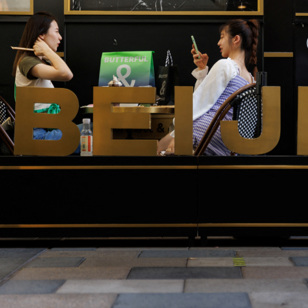 Two women relax in a cafe behind a sign with large letters spelling out Beijing, in a shopping area in Beijing, China, on July 14, 2022. 