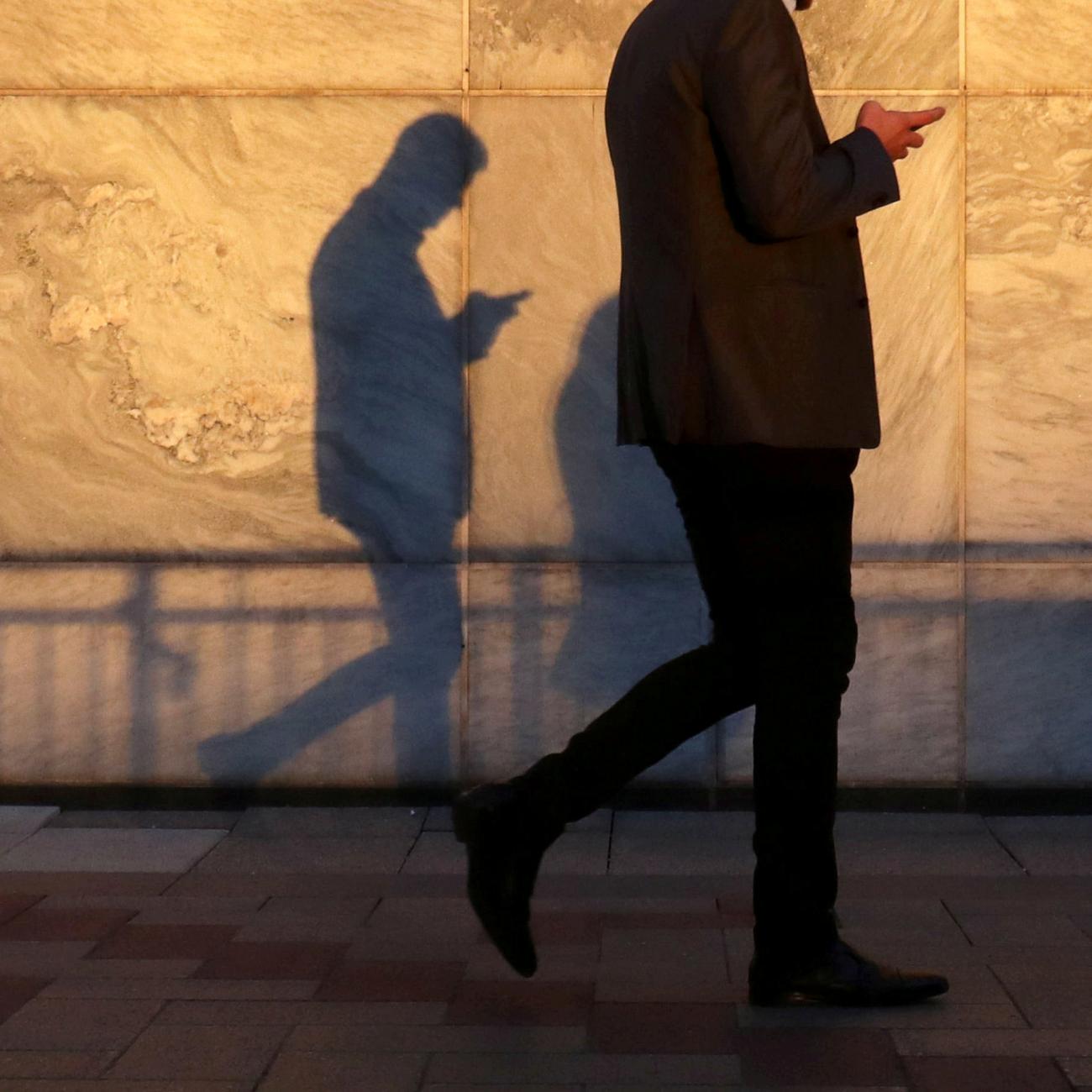 A man using a smart phone walks through London's Canary Wharf financial district in the warm yellow evening light, casting a shadow in London, United Kingdom, on September 28, 2018.