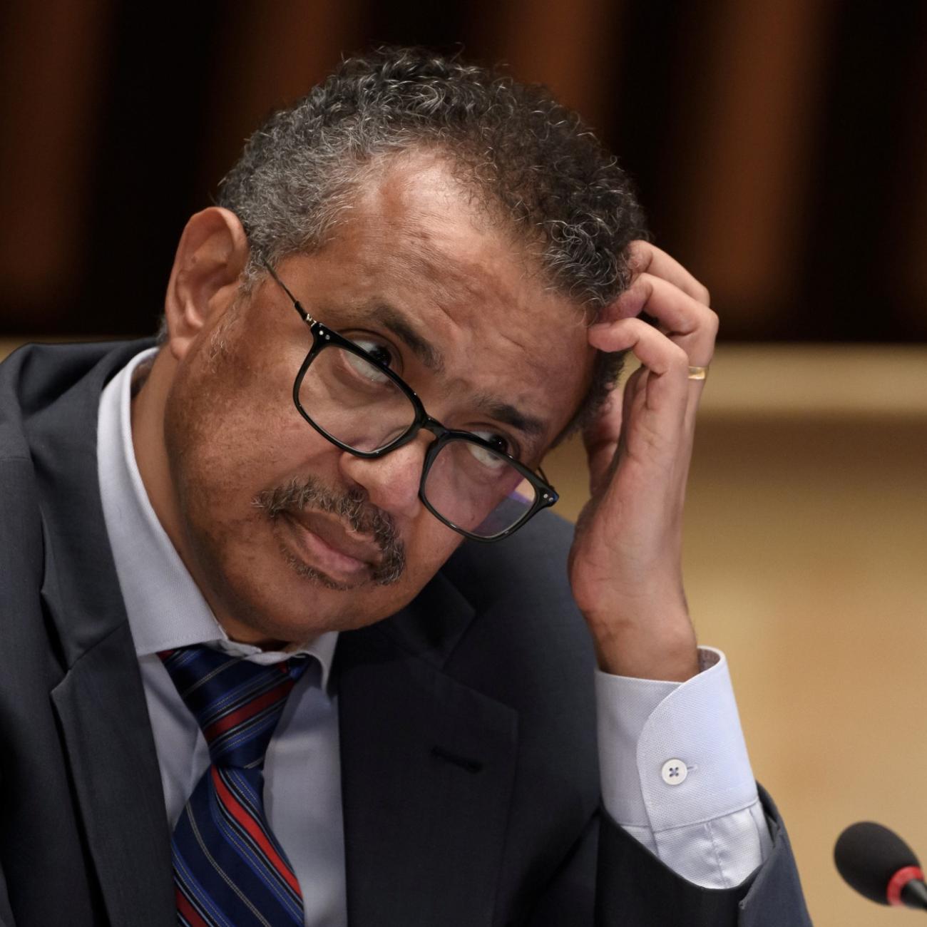 WHO Director-General Tedros Adhanom Ghebreyesus scratches his head with a quizzical expression while seated at a wooden podium 