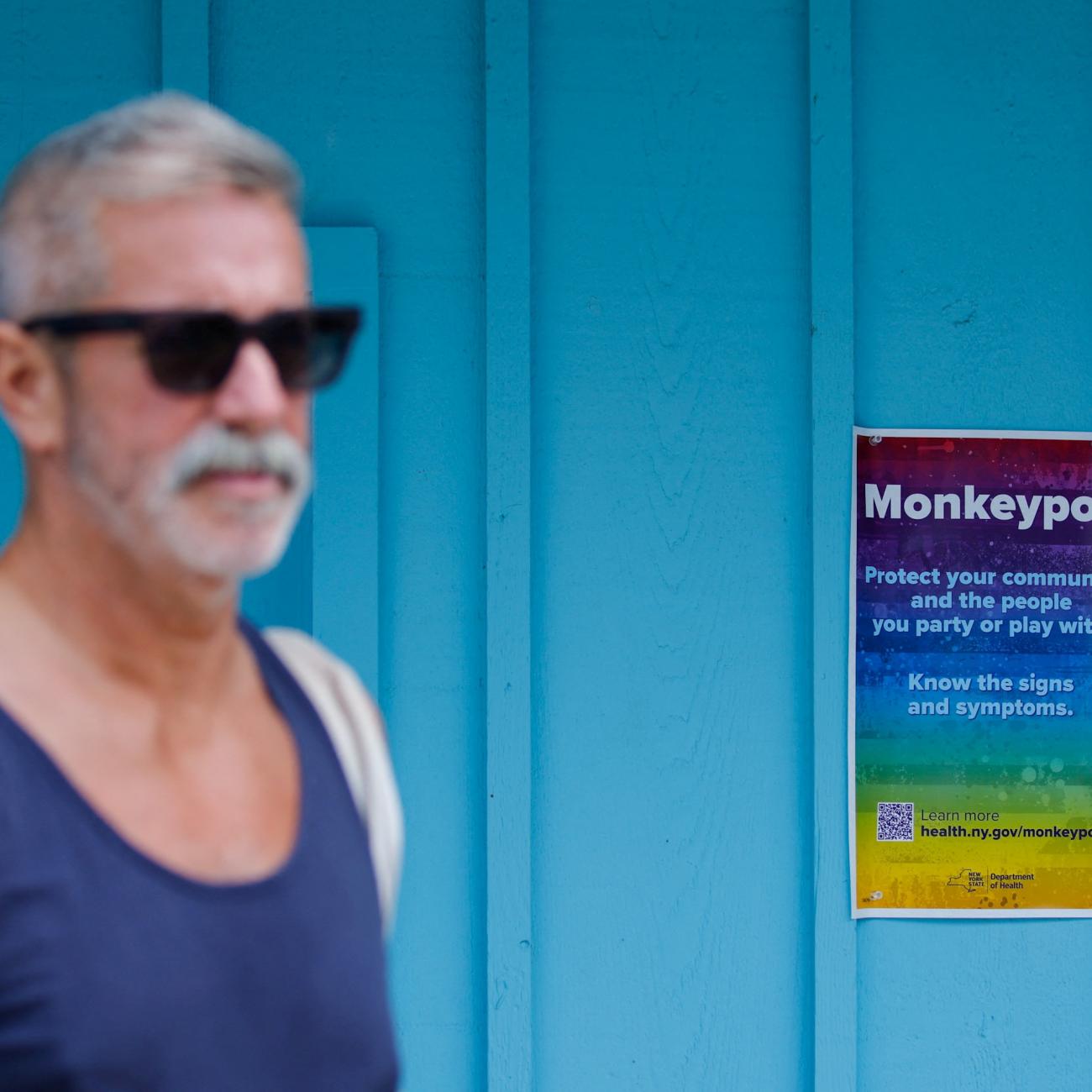 A grey-haired bearded man in a blue tank top and sunglasses walks past a turquoise wall with a rainbow poster that reads "Protect your community and the people you party or play with. Know the signs and symptoms" placed there by the New York Department of Health