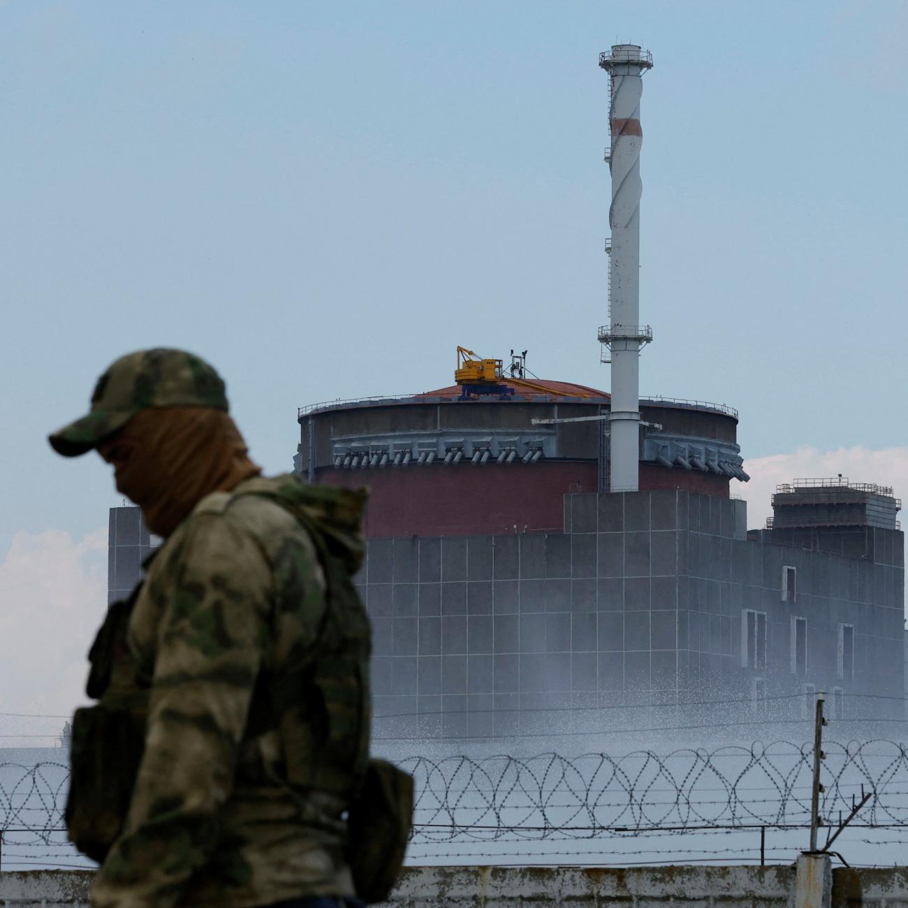 A Russian soldier in a camouflage uniform bearing the Russian flag and a cloth face covering stands guard near the Zaporizhzhia Nuclear Power Plant whose towers stand surrounded by a fog and a barbed wire against a blue sky. 