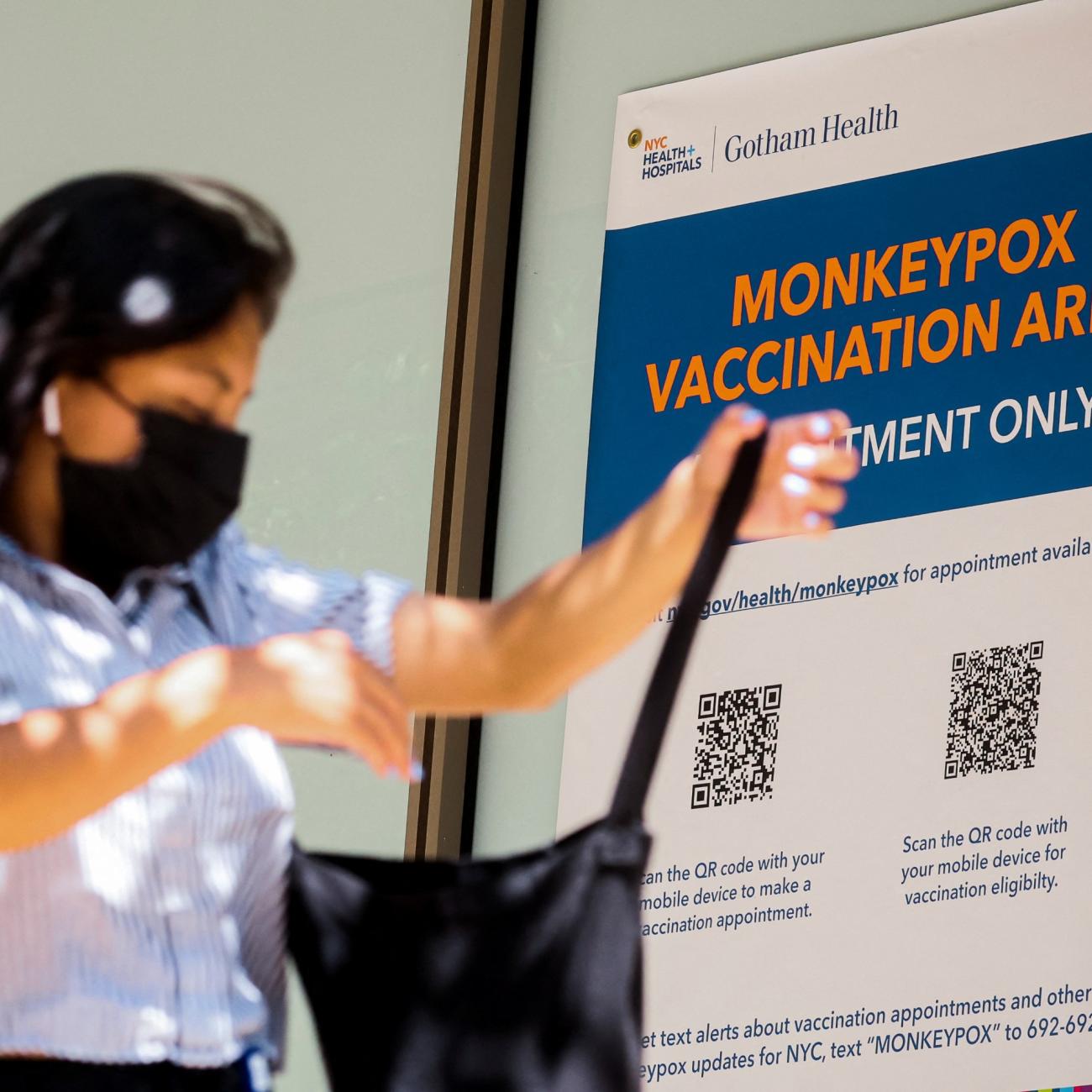 A woman with long dark hair in a light blue blouse and black mask searches for something in a black tote bag as she walks past a blue sign with orange letters reading "monkeypox vaccination area"