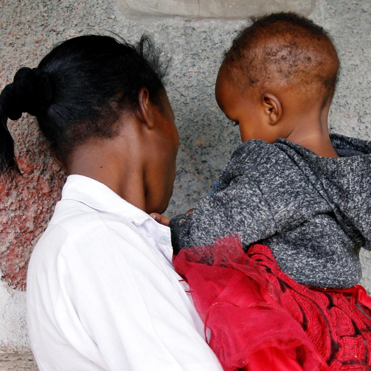 A young woman wearing a white shirt is photographed from behind holding her baby weraing a gray jumper