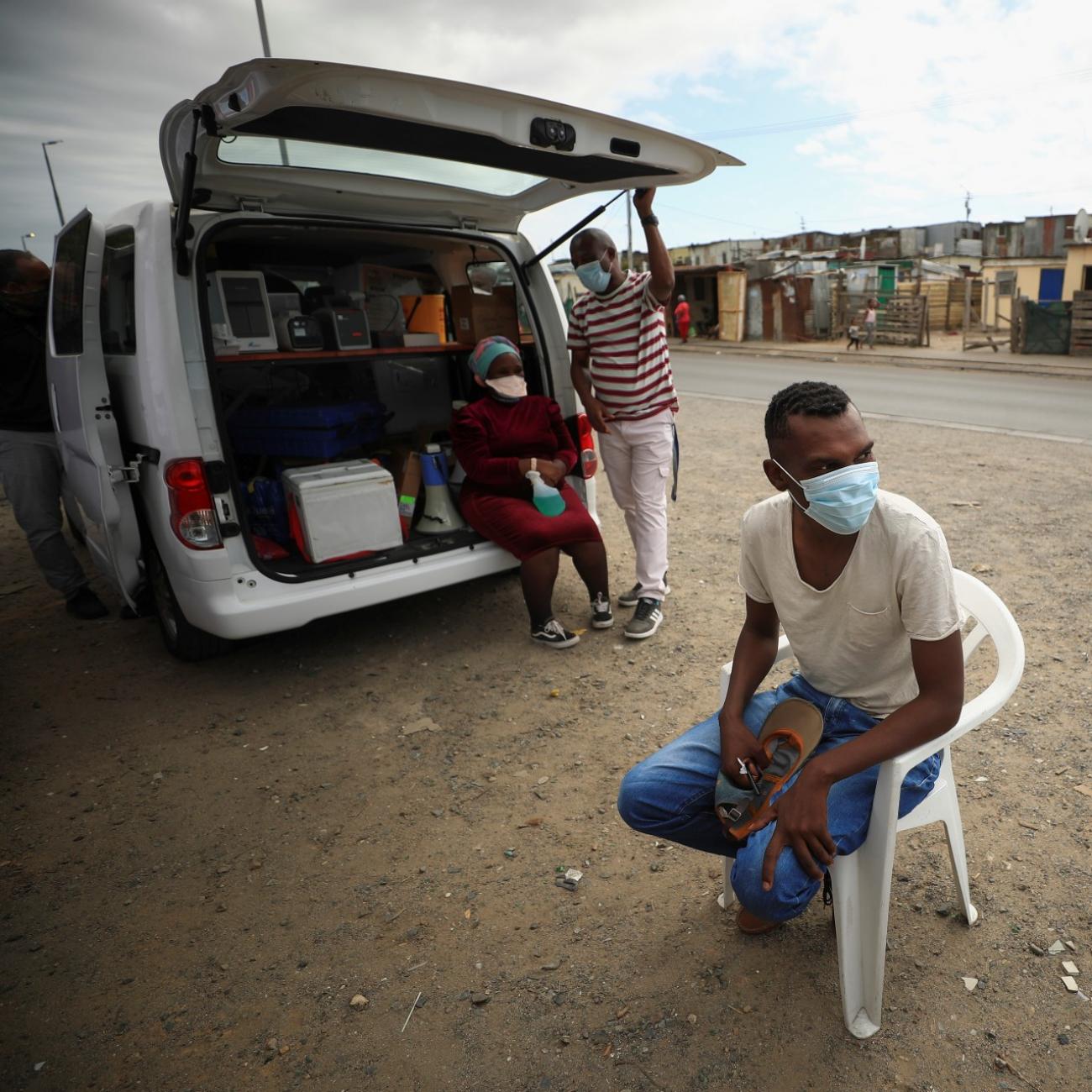 a young man wearing a white t-shirt, blue jeans, and a blue surgical face mask, sits on a plastic chair outside a mobile TB testing clinic