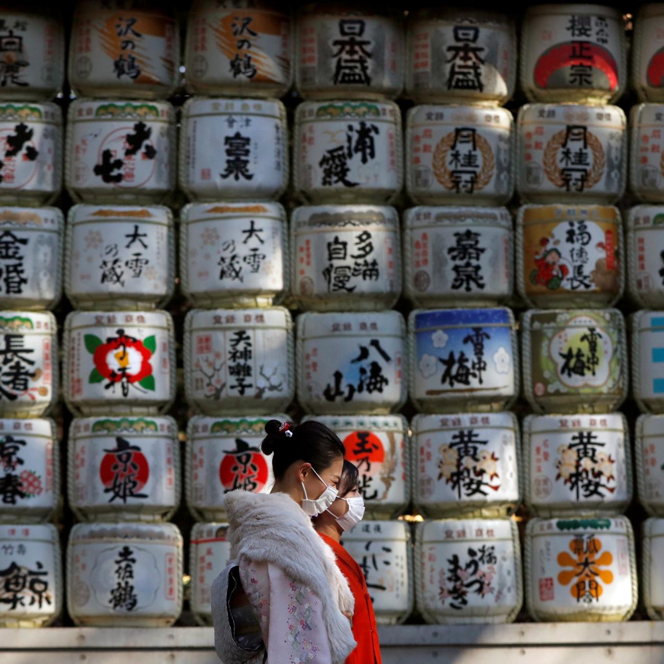 Kimono-clad women wearing protective face masks walk in front of white Japanese Sake barrels stacked in a grid decorated with colorful designs for the year-end and New-Year at Meiji Shrine