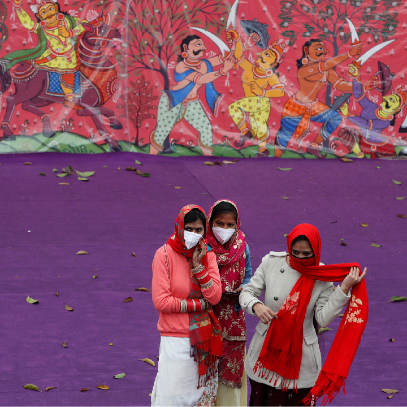 Women wearing protective face masks and colorful red and white saris stand in front of a purple wall and a color mural as they watch a dress rehearsal for the Republic Day parade, in New Delhi, India, on January 23, 2022. REUTERS/Adnan Abidi
