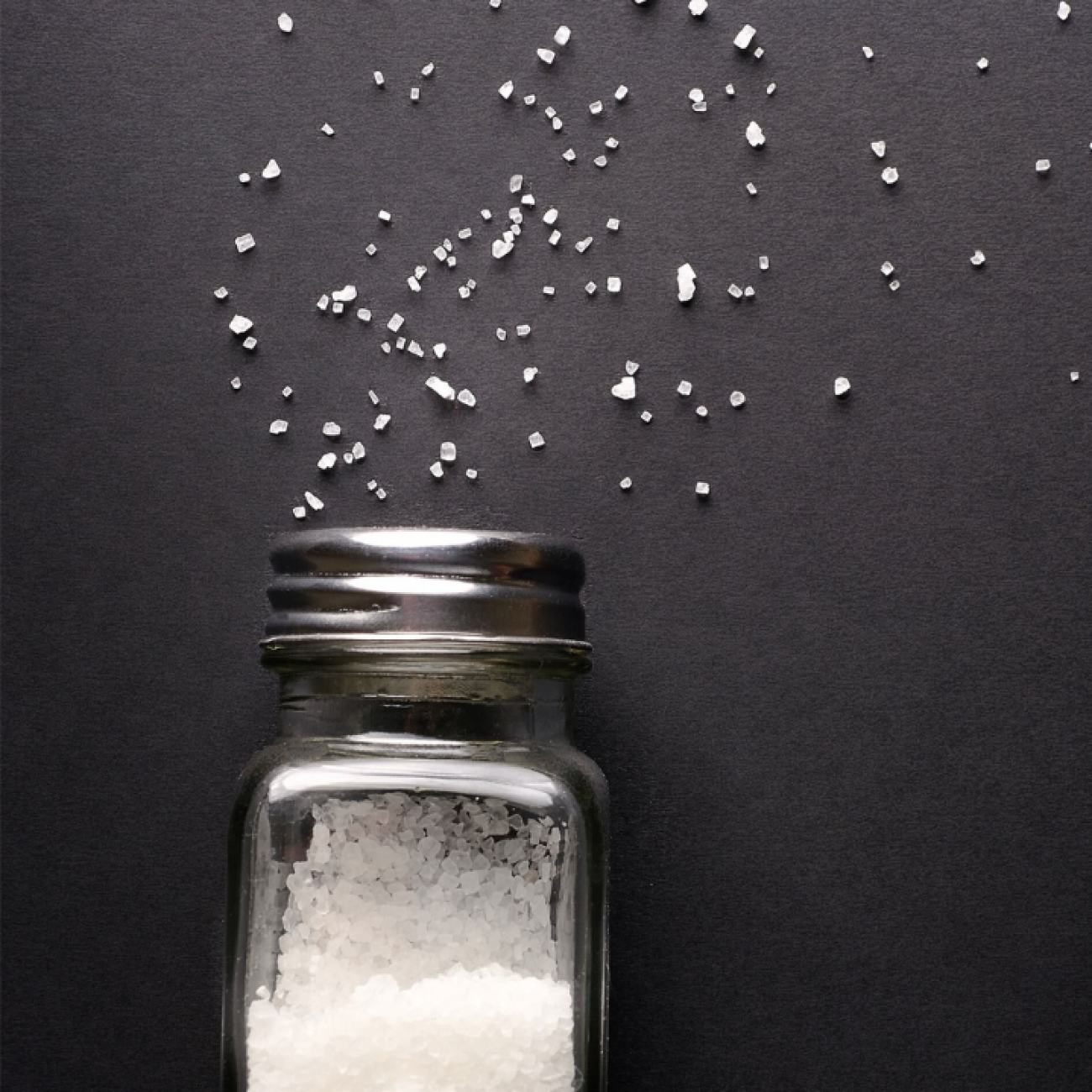 Natural, organic white sea salt pours from a fallen salt shaker onto a black table. 