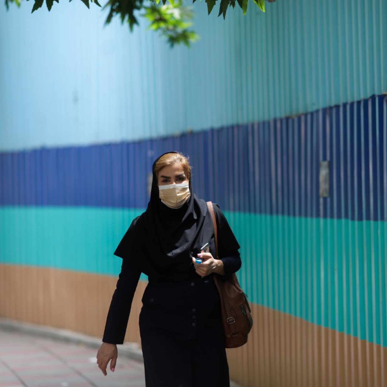 An woman walks along a street in Tehran, Iran, on May 26, 2021. Behind her stretches a wall with thick colorful horizontal stripes in shadess of blue and aqua.