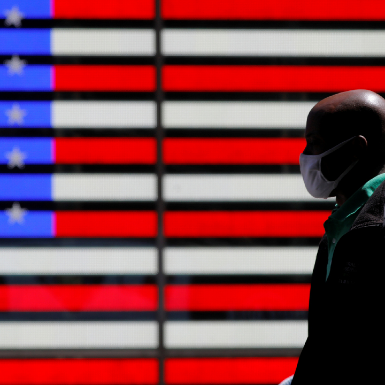 A giant red, white, and blue lit-up U.S. flag can be seen behind a person wearing a mask while walking in Times Square after the CDC announced new guidelines regarding outdoor mask wearing and vaccination during the COVID outbreak in New York City, New York, on April 27, 2021. 