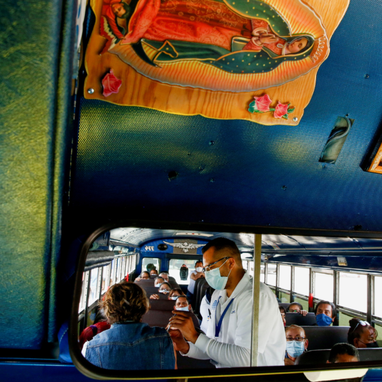 A rearview mirroron a bus reflects the image of assembly factory employees sitting in the bus waiting to receive COVID-19 vaccines. A health worker in a white medical coat is giving a vaccine to one of the women. Photo taken in Ciudad Juarez, Mexico, on May 24, 2021. 