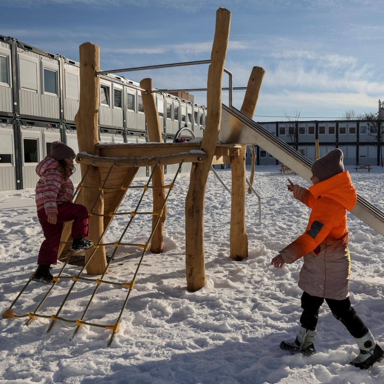 a child in an orange puffer jacket pushes their friend on a wooden swing in the snow