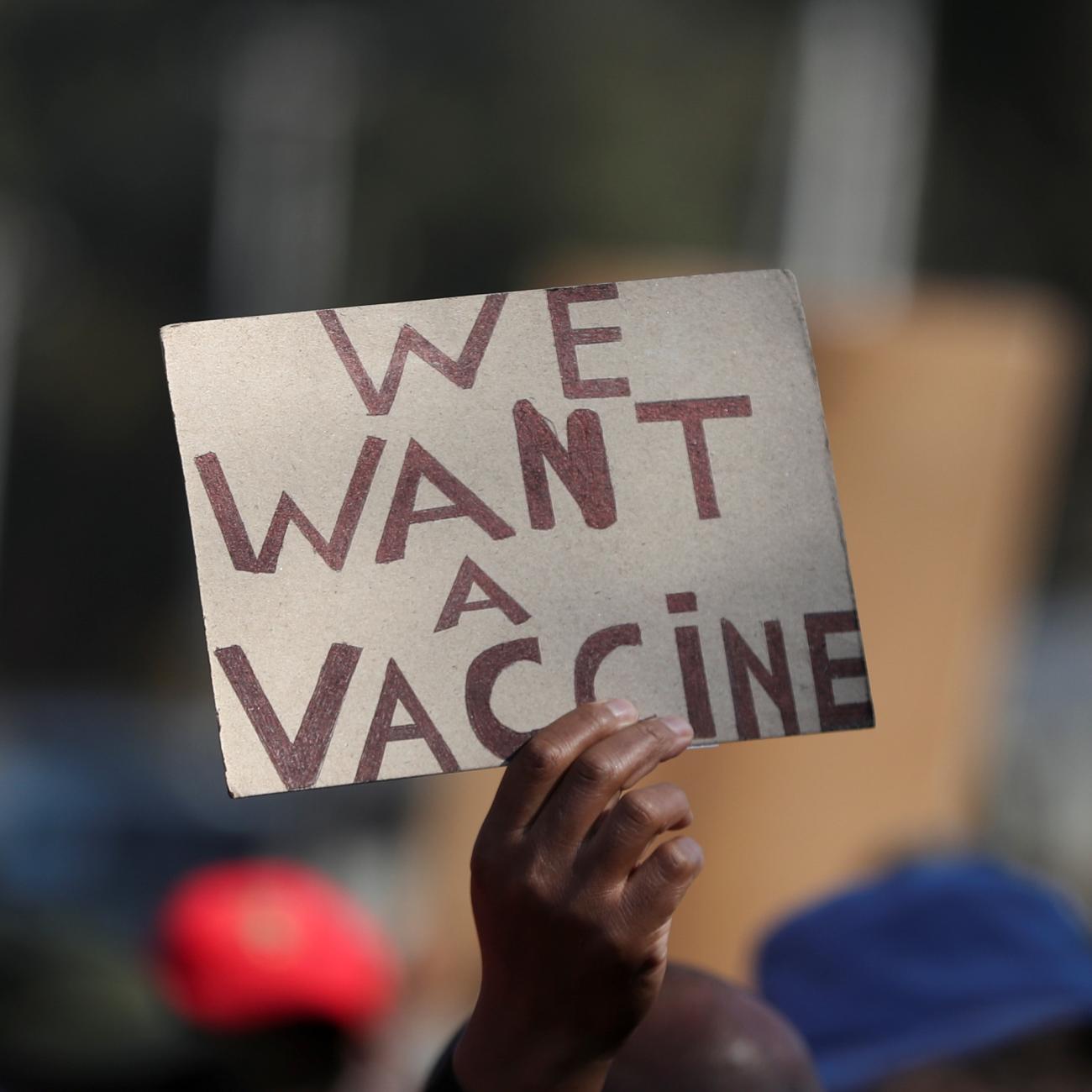 Hand holding up a sign that reads "We Want a Vaccine"