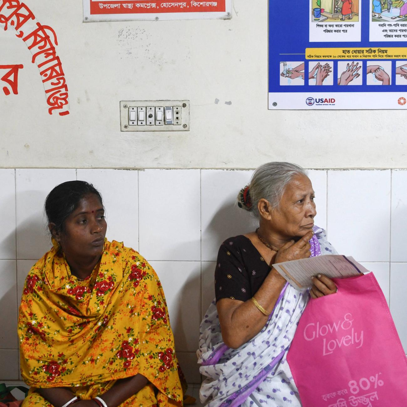 Patients wait for treatment as a USAID-funded health awareness poster hangs behind them, at the Hossainpur Upazila Health Complex, in Kishoreganj, Bangladesh.